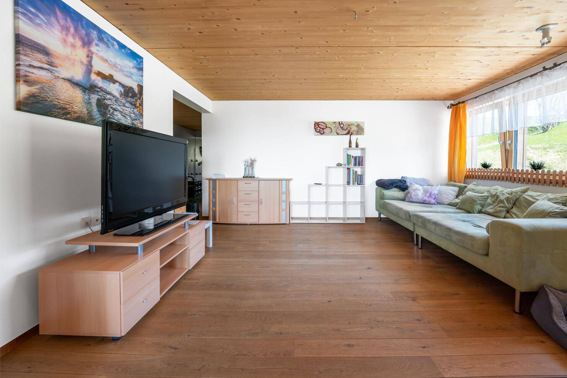 Helles Wohnzimmer mit großem Fernseher, hellgrünem Sofa und Holzdielenboden, mit Blick ins Grüne.