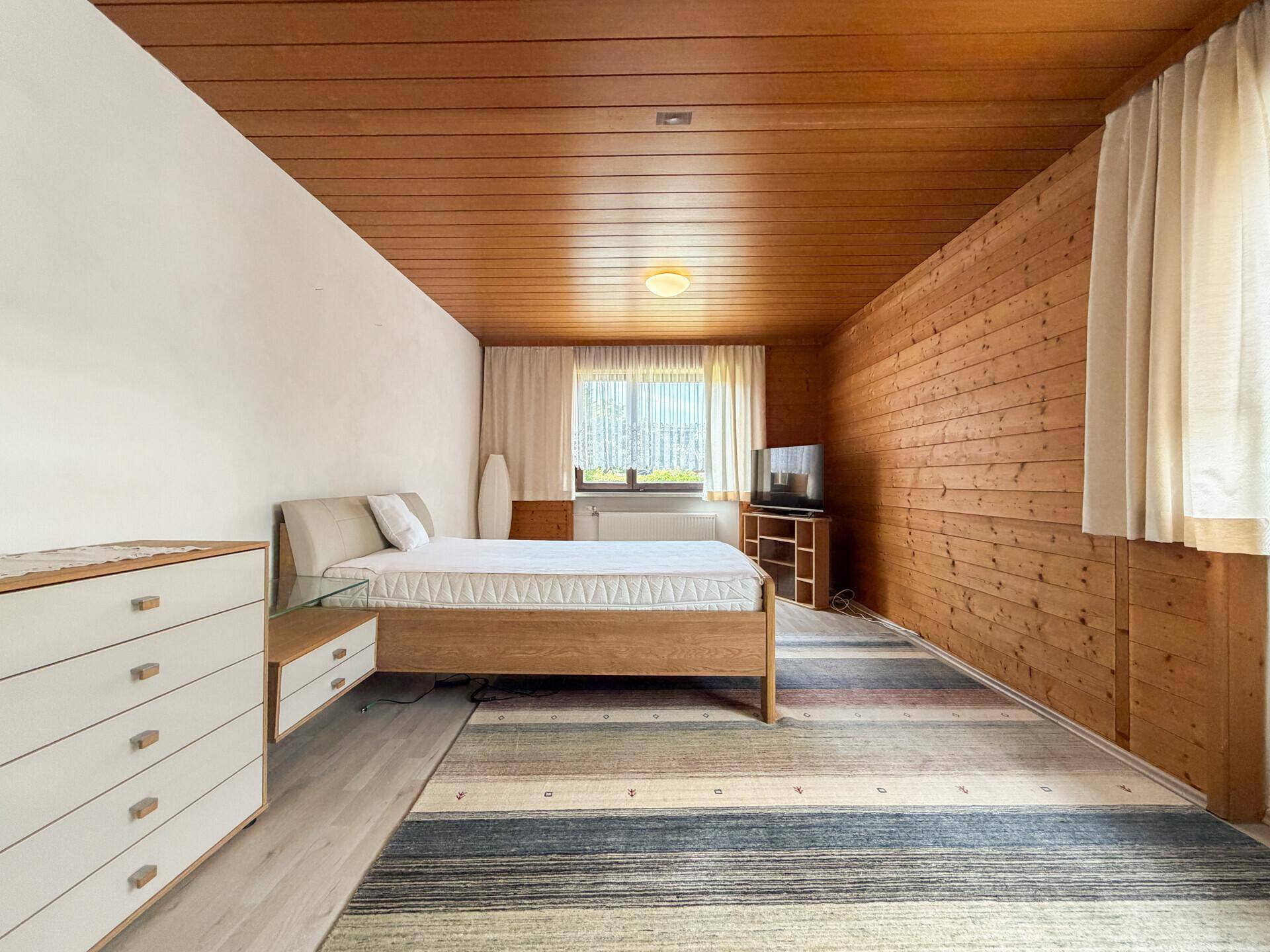 Helles Schlafzimmer mit Holzdecke, Holzbett und einem Teppich auf dem Boden.