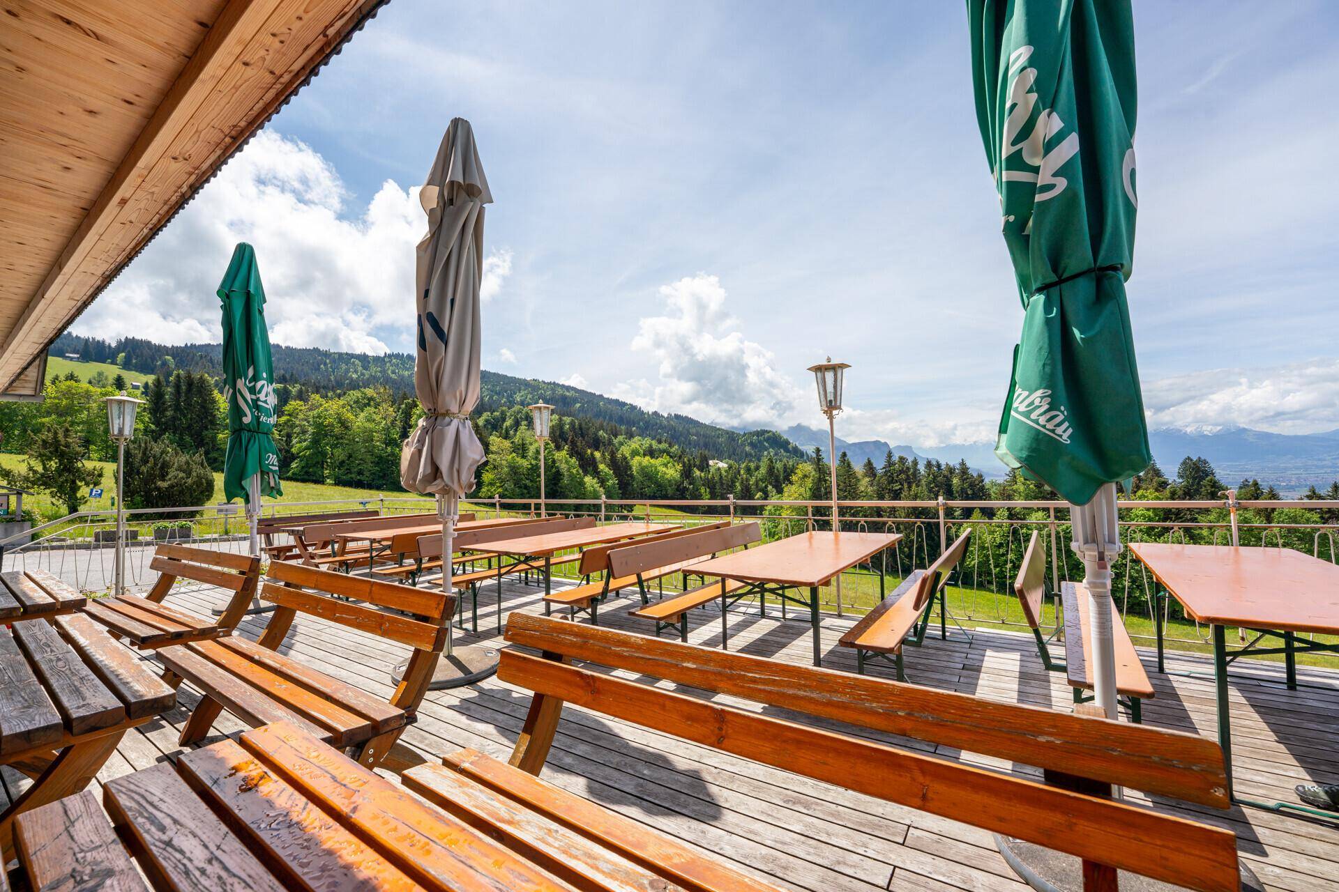 Große Holzterrasse mit Biertischen und Sonnenschirmen, umgeben von grünen Hügeln und Bergen im Hintergrund.