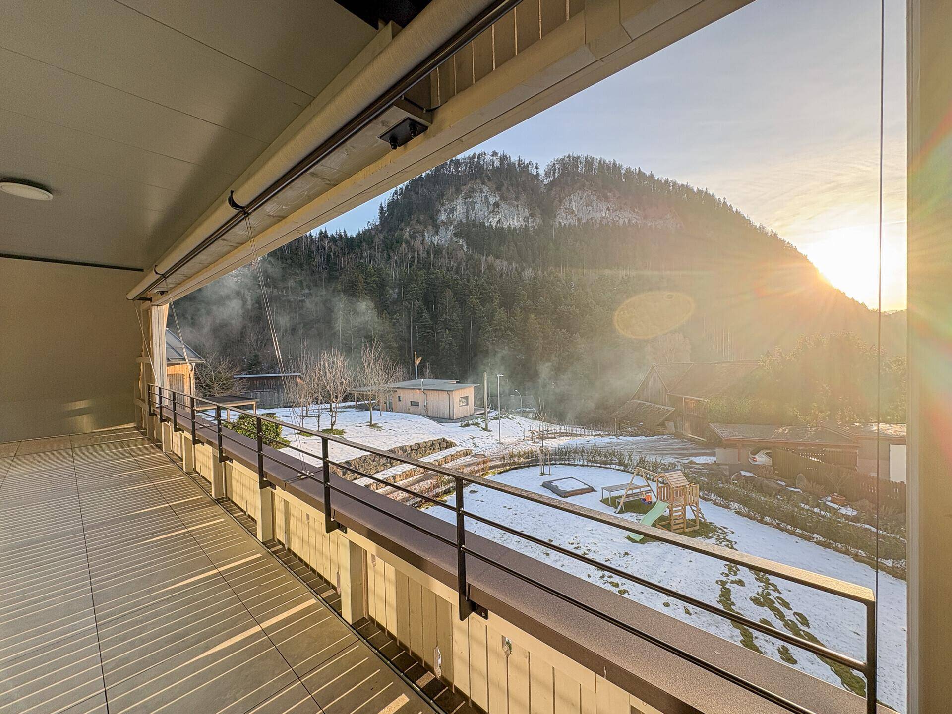 Sonniger Balkon mit Panoramablick auf die Berge und den Spielplatz im verschneiten Garten.