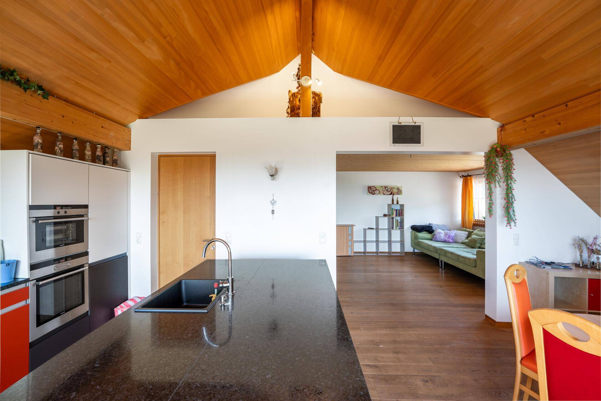 Offener Wohnbereich mit Kücheninsel im Vordergrund und gemütlichem Wohnzimmer im Hintergrund, unter einer Holzdecke.