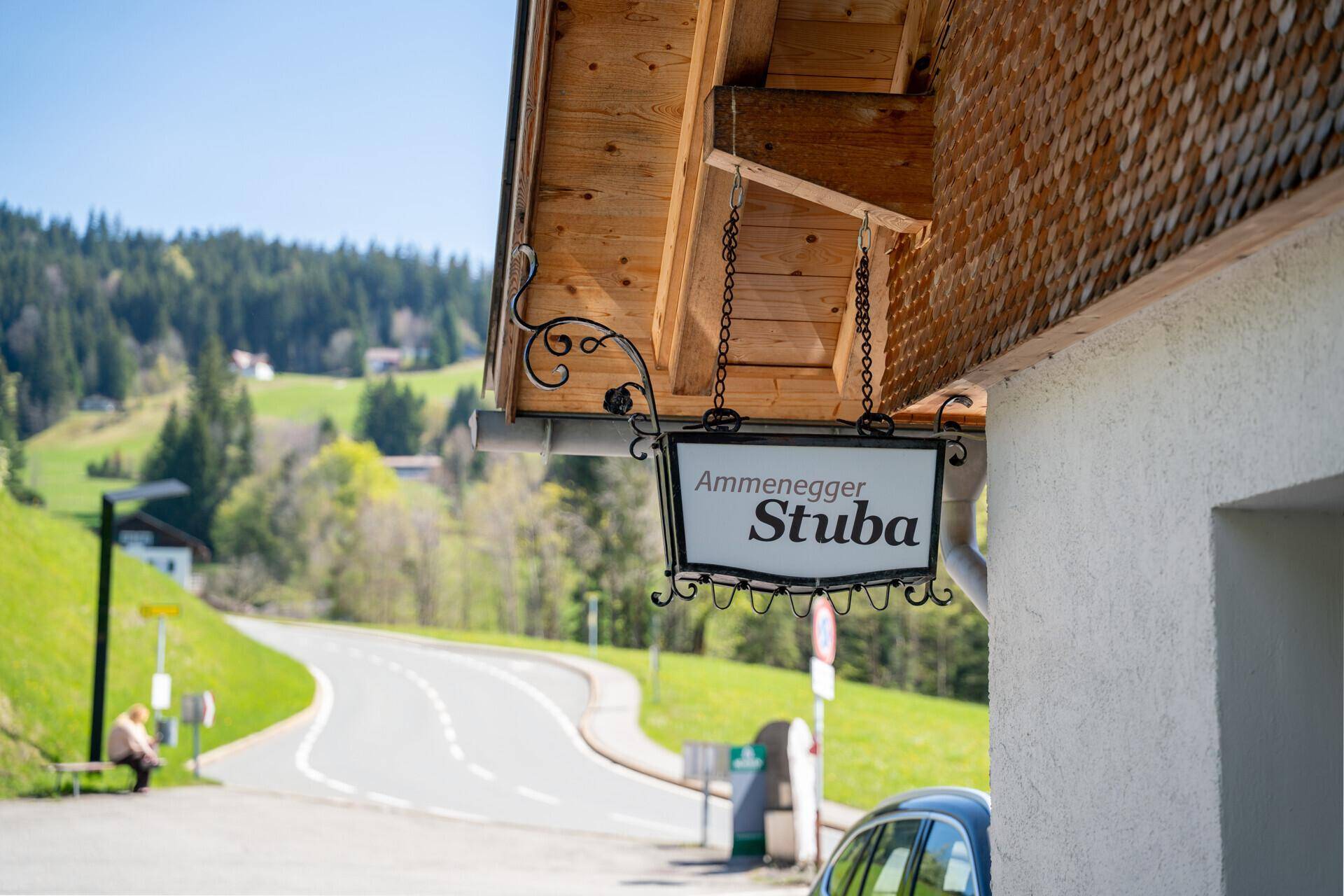 Ein traditionelles Schild mit dem Namen "Ammenegger Stuba" hängt unter einem Holzdachvorsprung an einem sonnigen Tag.
