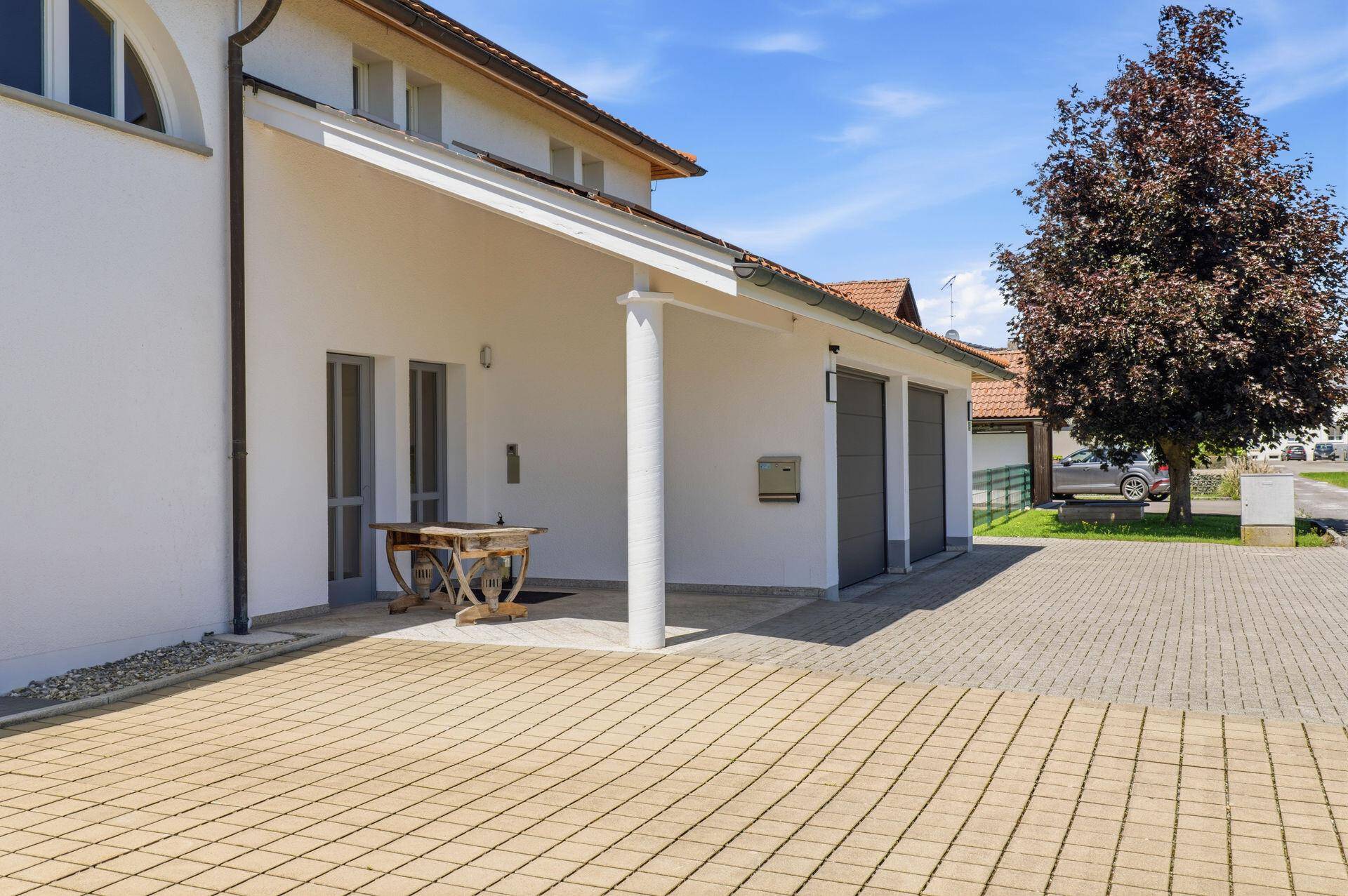 Eingangsbereich mit überdachtem Carport und gepflasterter Auffahrt, umgeben von Grün.