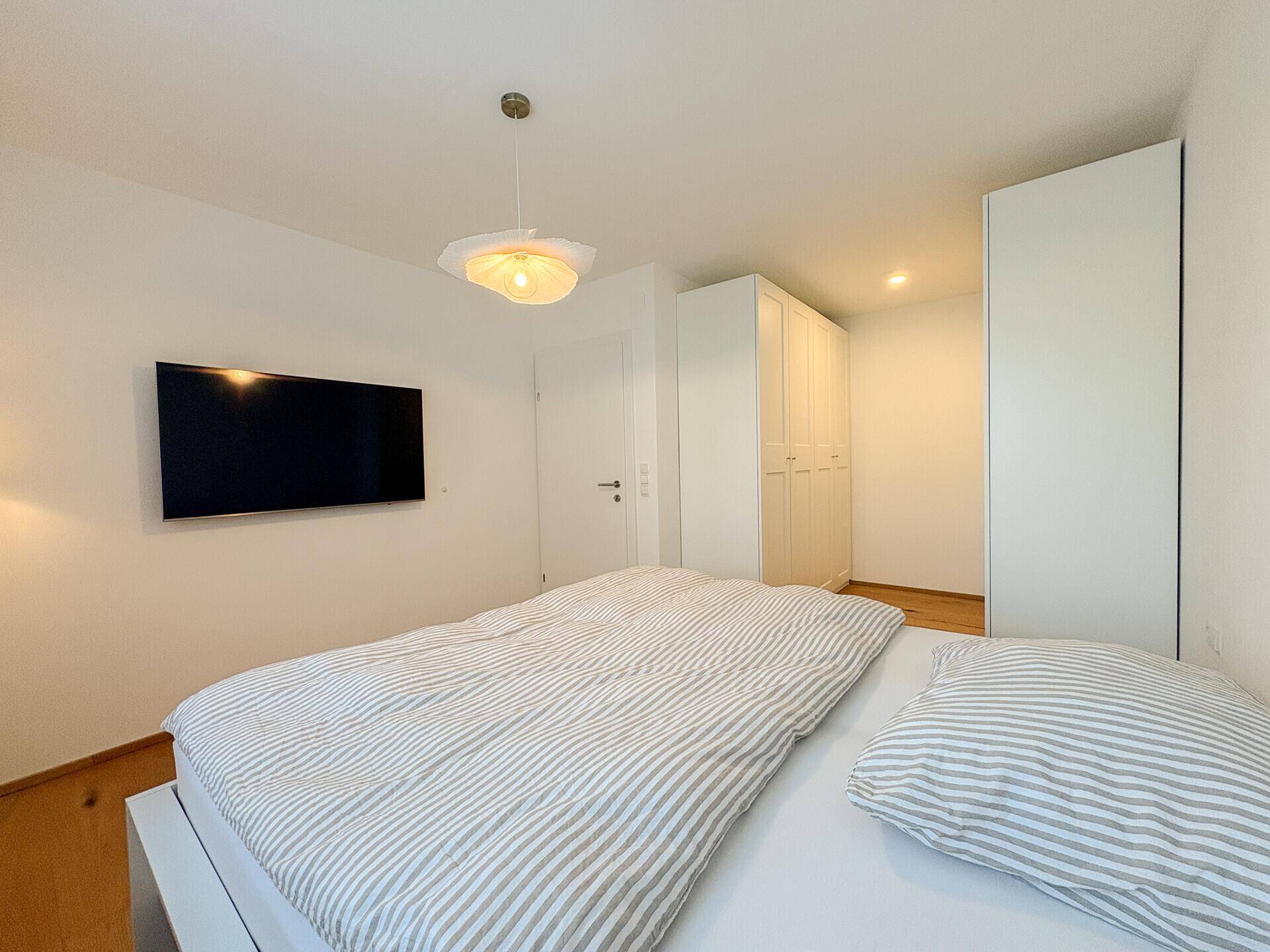Gemütliches Schlafzimmer mit Holzboden, weißem Bett und Wandfernseher.