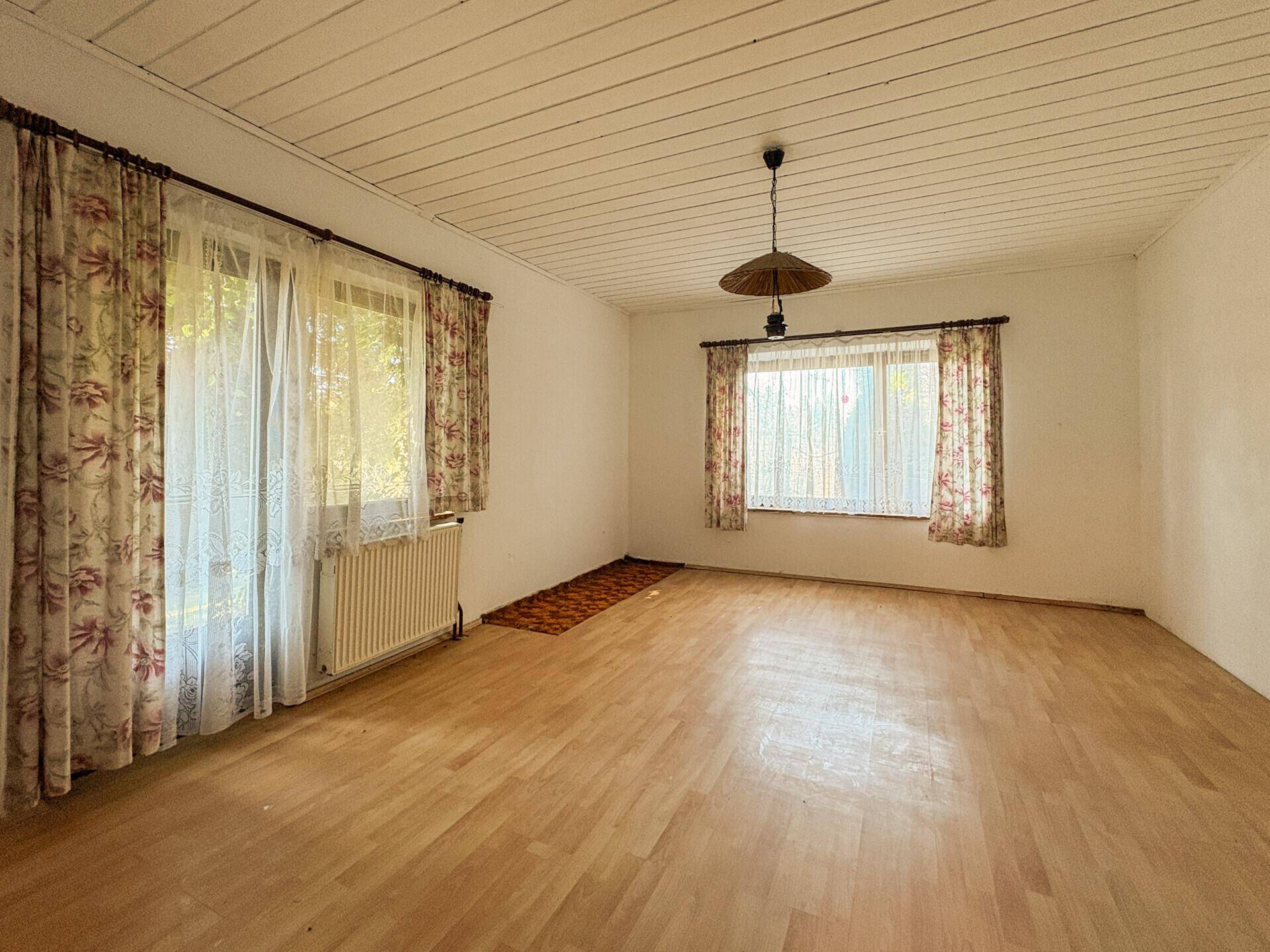Helles Wohnzimmer mit Holzfußboden, weißer Holzdecke und zwei Fenstern mit Vorhängen.