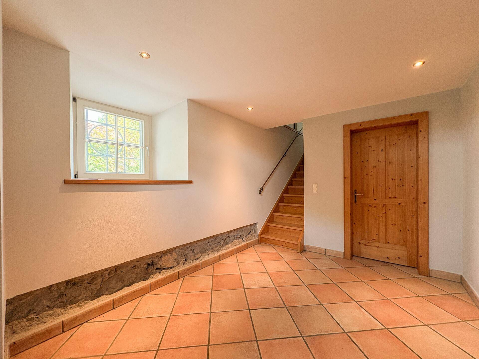 Heller Flur mit Terrakottafliesen, Holztreppe und kleinem Fenster.