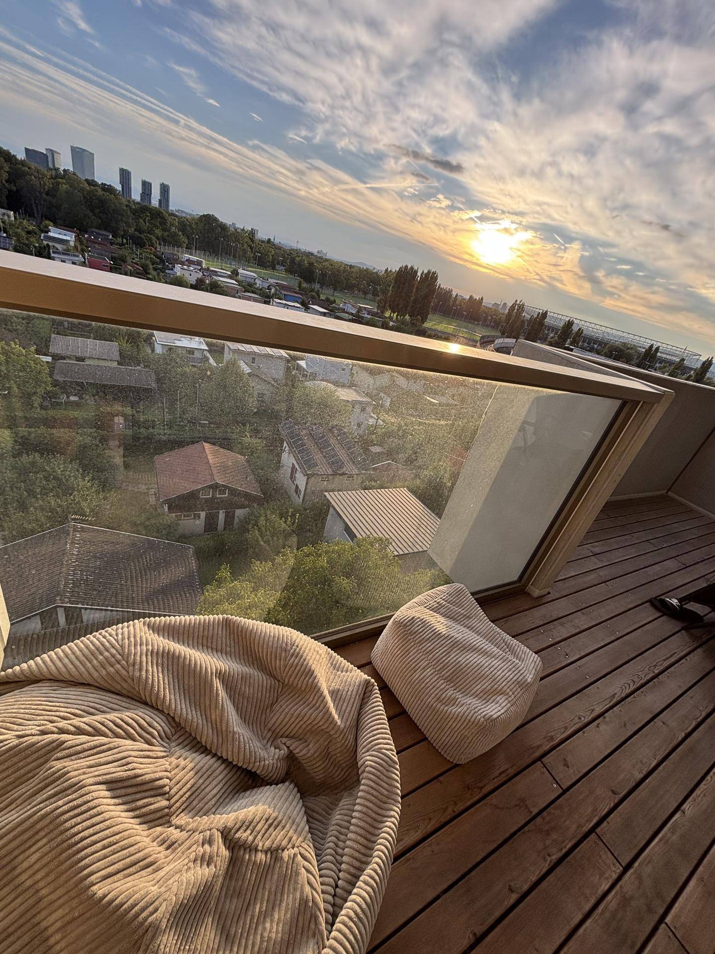 Gemütlicher Balkon mit Holzdeck und Sitzsäcken, bietet einen weiten Blick über die Stadt bei Sonnenuntergang.