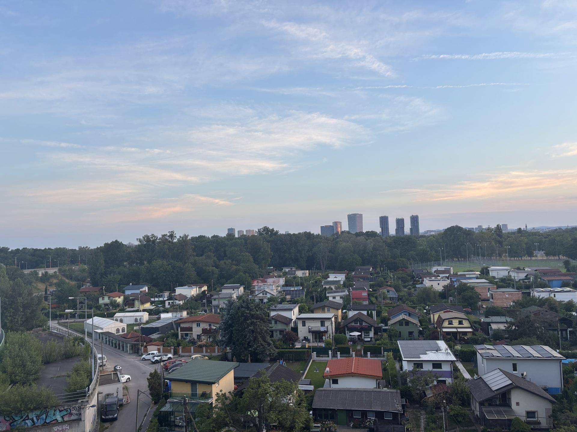 Weitläufiger Blick über eine Vorstadtlandschaft mit Häusern, Bäumen und Hochhäusern am Horizont.