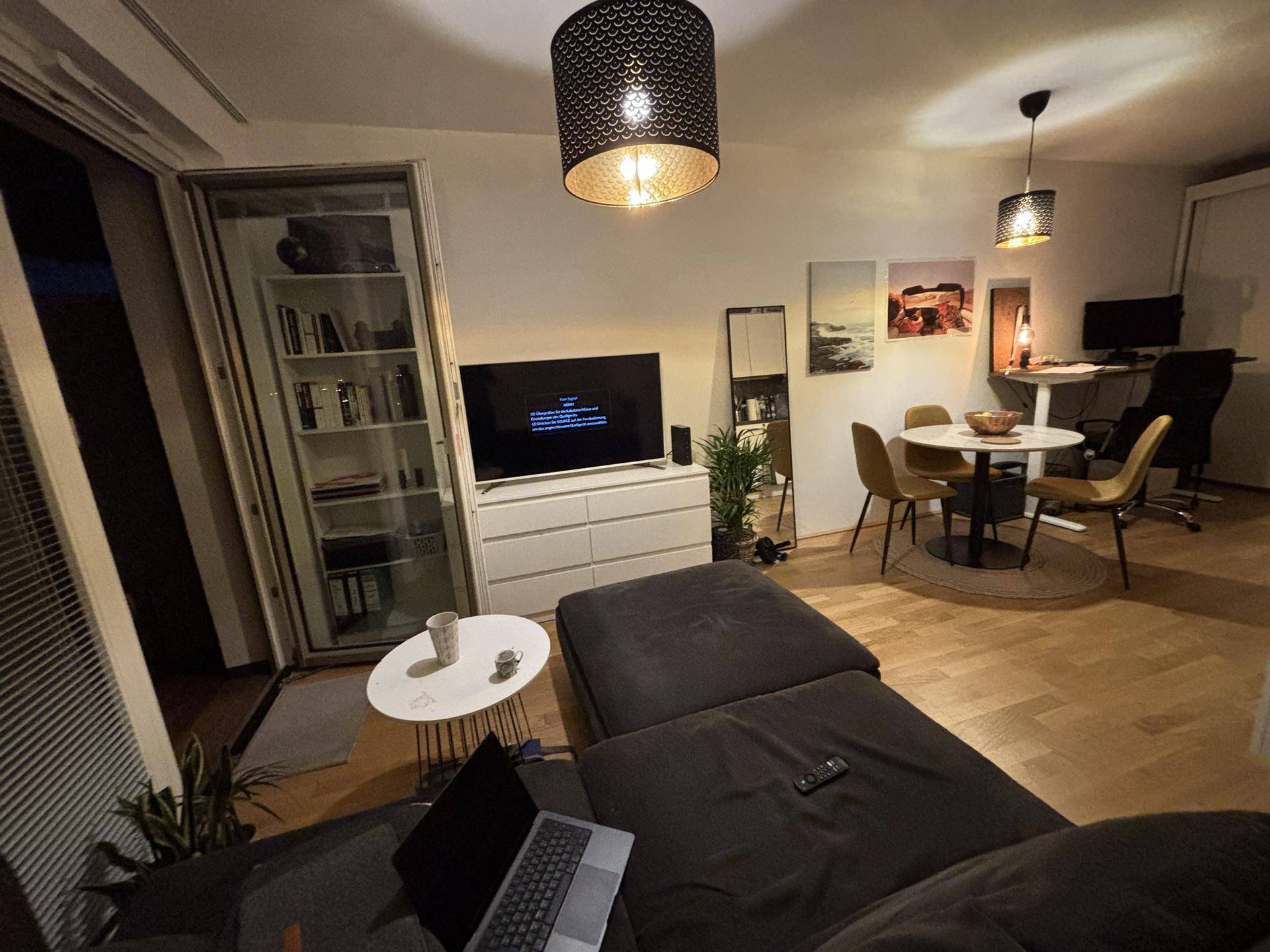 Heller Wohn- und Essbereich mit modernem Sofa, Fernseher und einem funktionalen Arbeitsbereich.