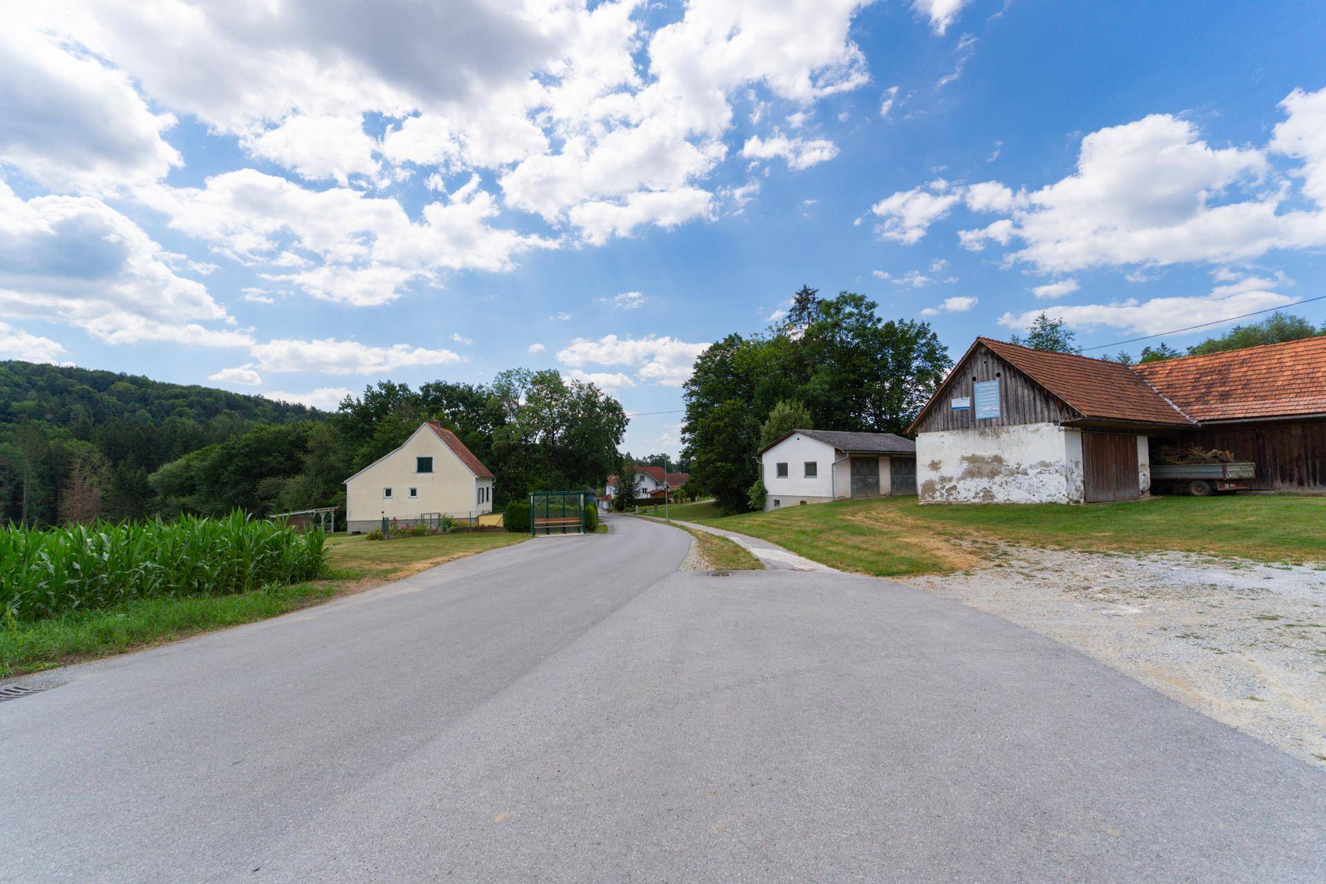 Ländliche Straße führt an einem alten Haus vorbei, mit weiteren Gebäuden und Wald im Hintergrund.