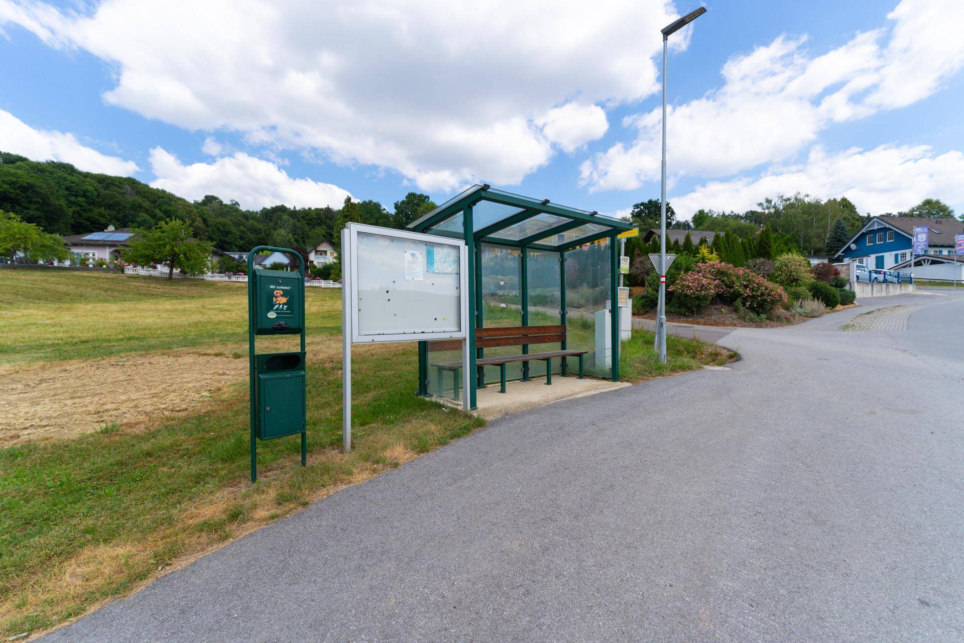 Bushaltestelle mit Sitzbank und Mülleimer an einer Landstraße, umgeben von Häusern und Natur.
