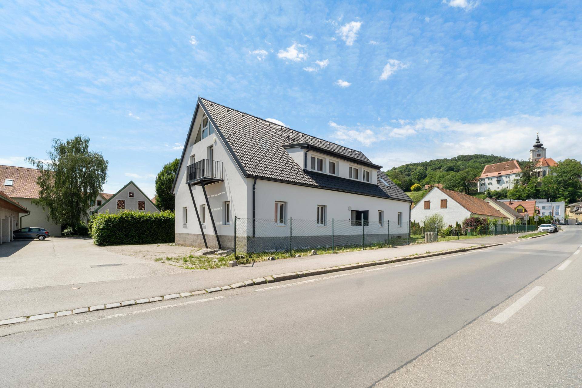 Modernes Haus mit Satteldach und Balkon, gelegen an einer Straße mit Blick auf eine Kirche.