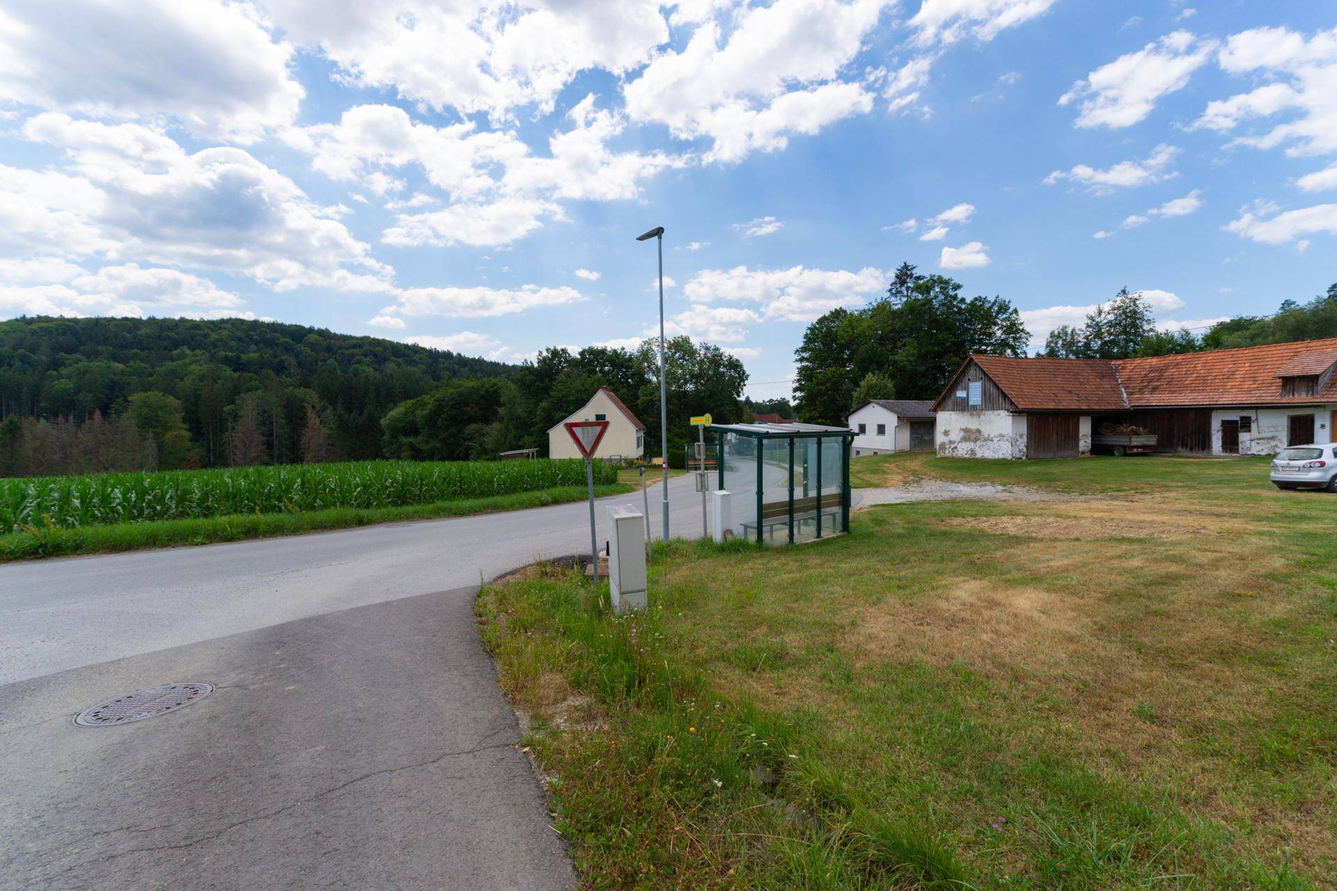 Ländliche Straße mit Bushaltestelle, Gebäuden und einem Maisfeld vor bewaldeten Hügeln.