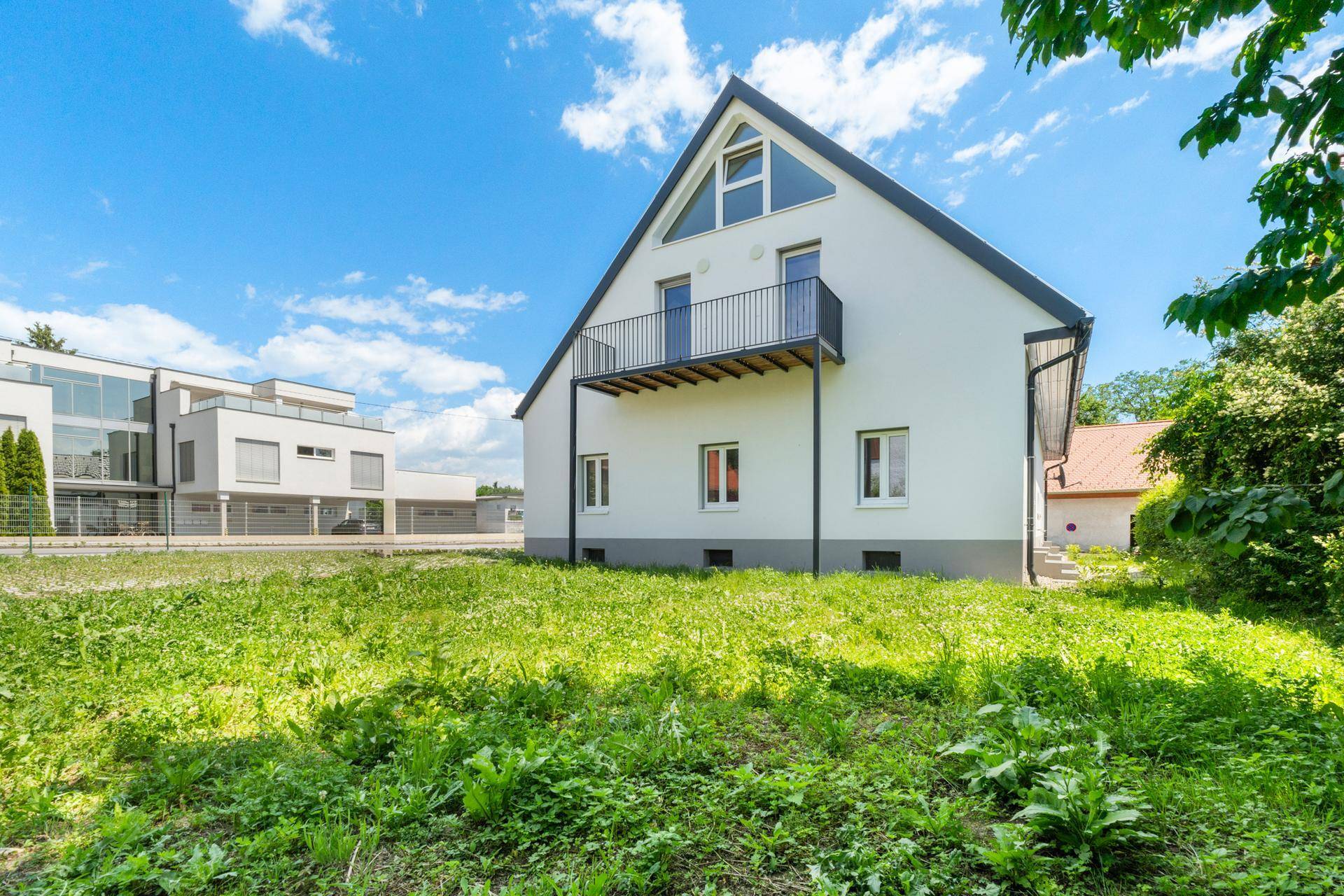 Seitenansicht des Hauses mit Rasenfläche und einem modernen Gebäude im Hintergrund.