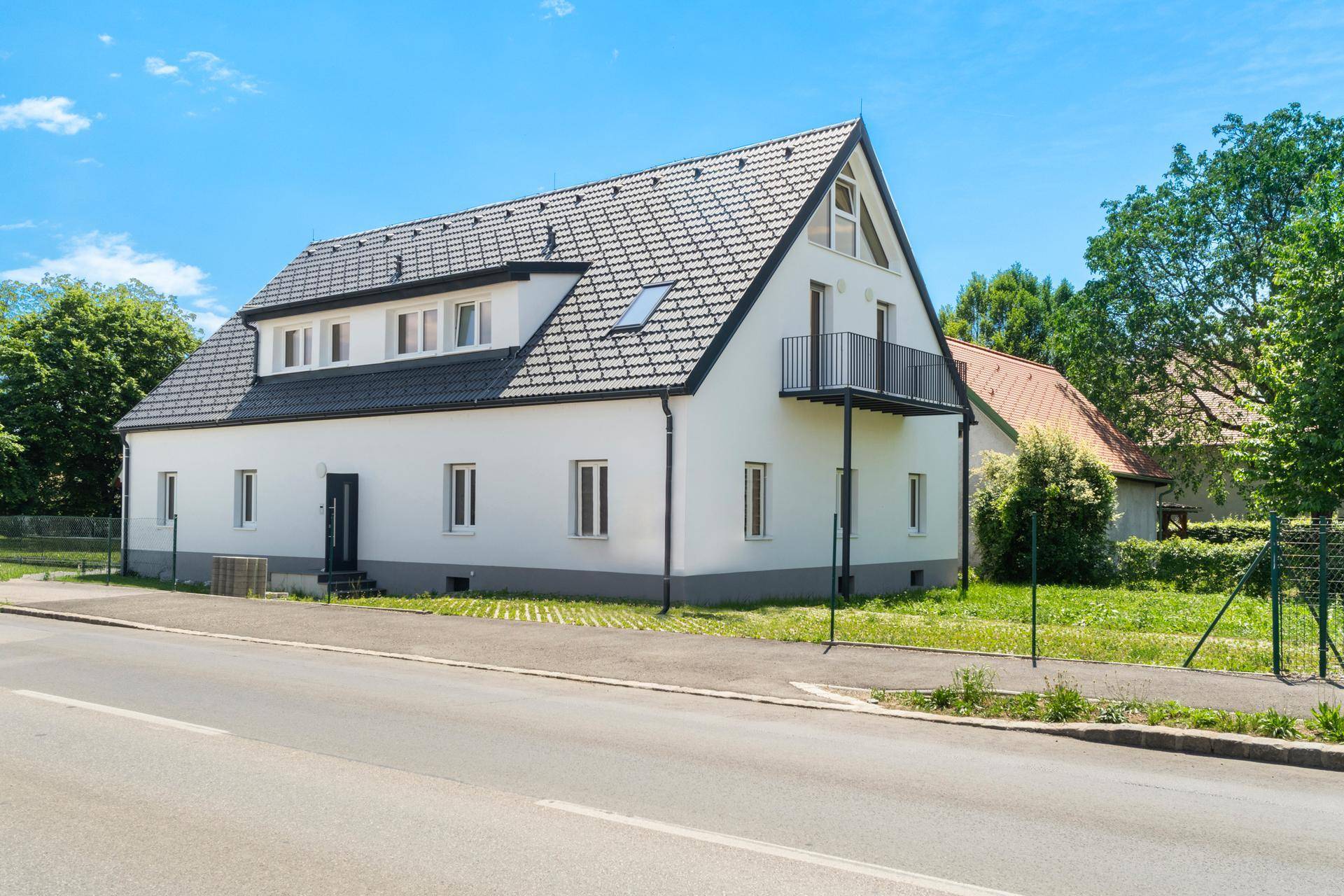 Seitenansicht des Hauses mit Balkon und gepflegter Umgebung unter blauem Himmel.