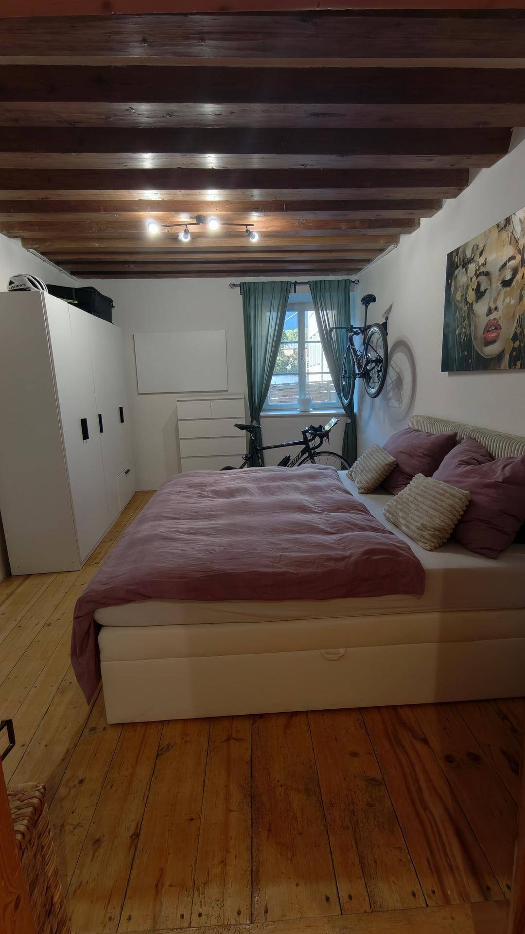 Schlafzimmer mit sichtbaren Holzbalken, Holzboden und einem Fenster mit Vorhängen.