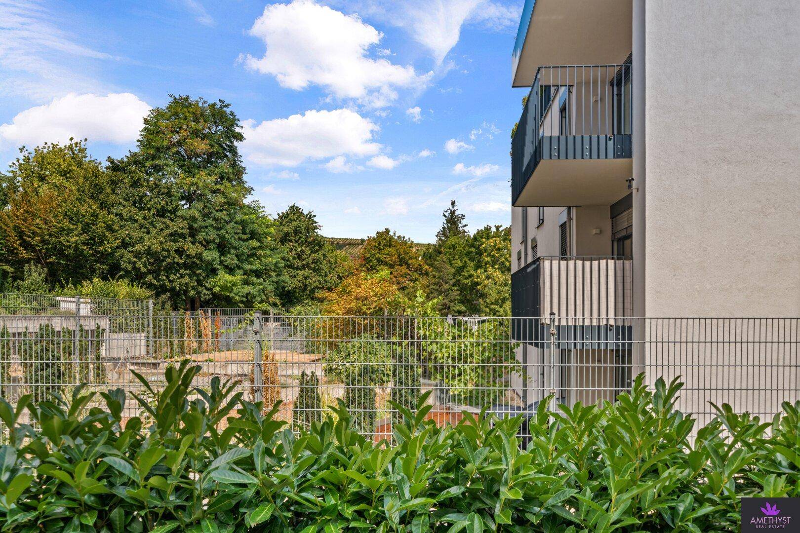 Blick ins Grüne mit Bäumen und einem Balkon im Hintergrund unter blauem Himmel.