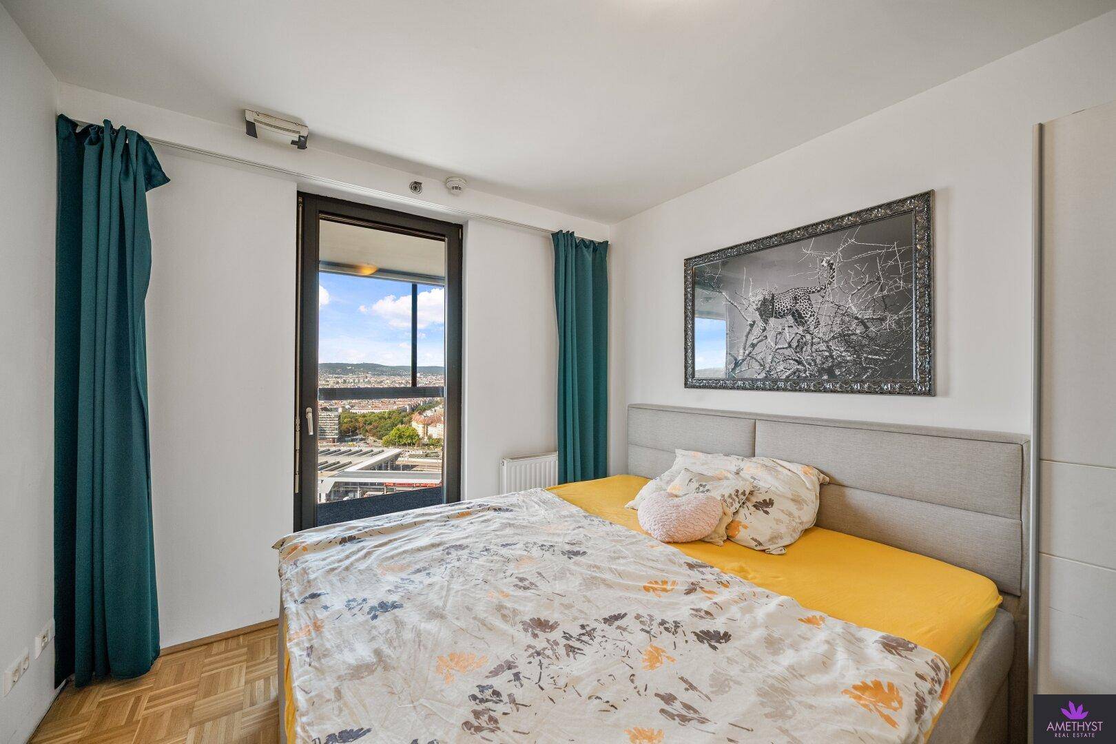 Schlafzimmer mit Doppelbett, Parkettboden und Zugang zu einem Balkon mit Stadtblick.