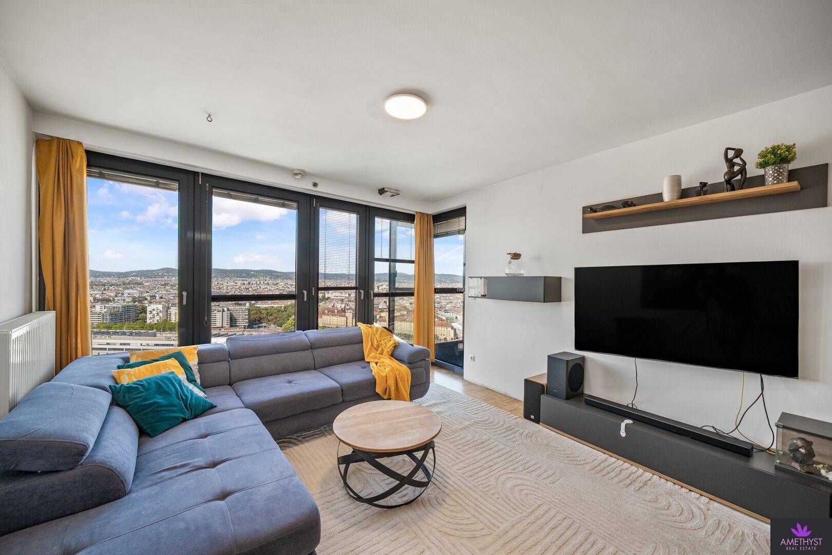 Helles Wohnzimmer mit bequemer Couch, Fernseher und großen Fenstern mit urbanem Ausblick.