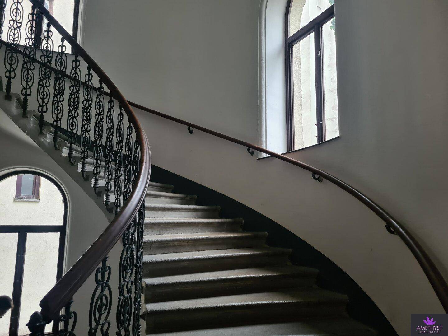 Elegantes geschwungenes Treppenhaus mit Holzhandlauf, schmiedeeisernem Geländer und hohen Bogenfenstern.