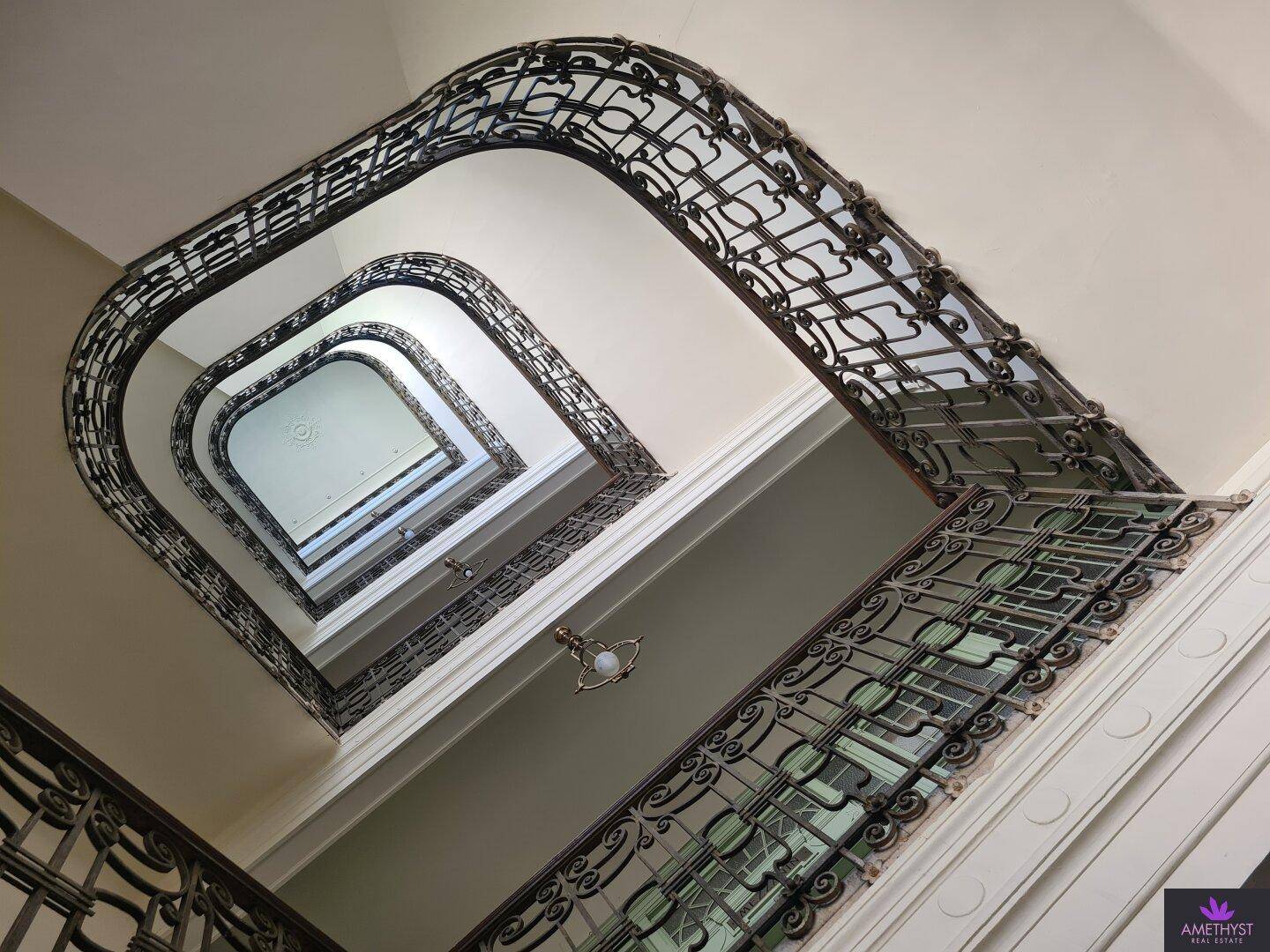 Blick von unten in ein historisches Treppenhaus mit kunstvollen schmiedeeisernen Geländern und hohen Decken.