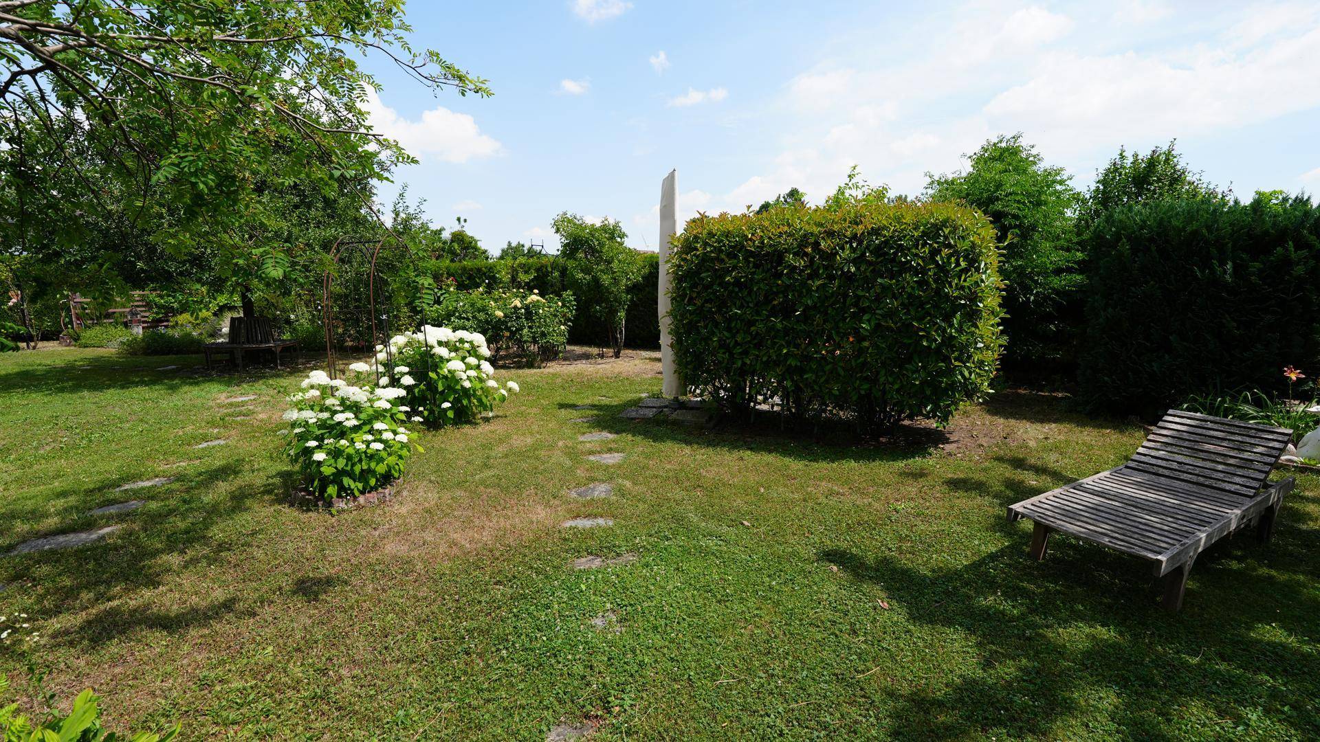 Großer, sonniger Garten mit Rasenfläche, blühenden Sträuchern und einer Holzliege zum Entspannen.
