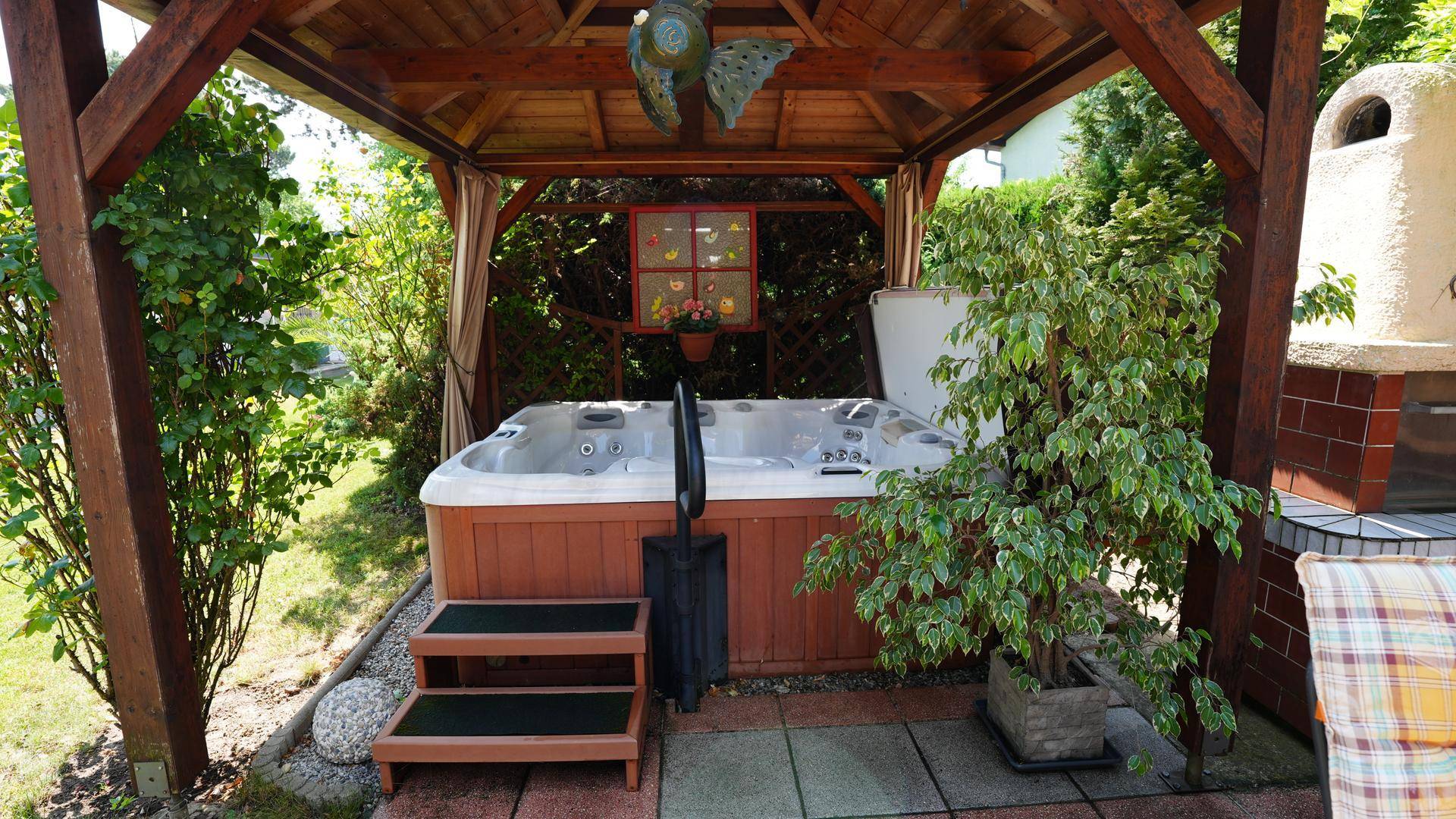 Gemütlicher Whirlpoolbereich unter einem Pavillon mit Außengrill und Pflanzen im Garten.
