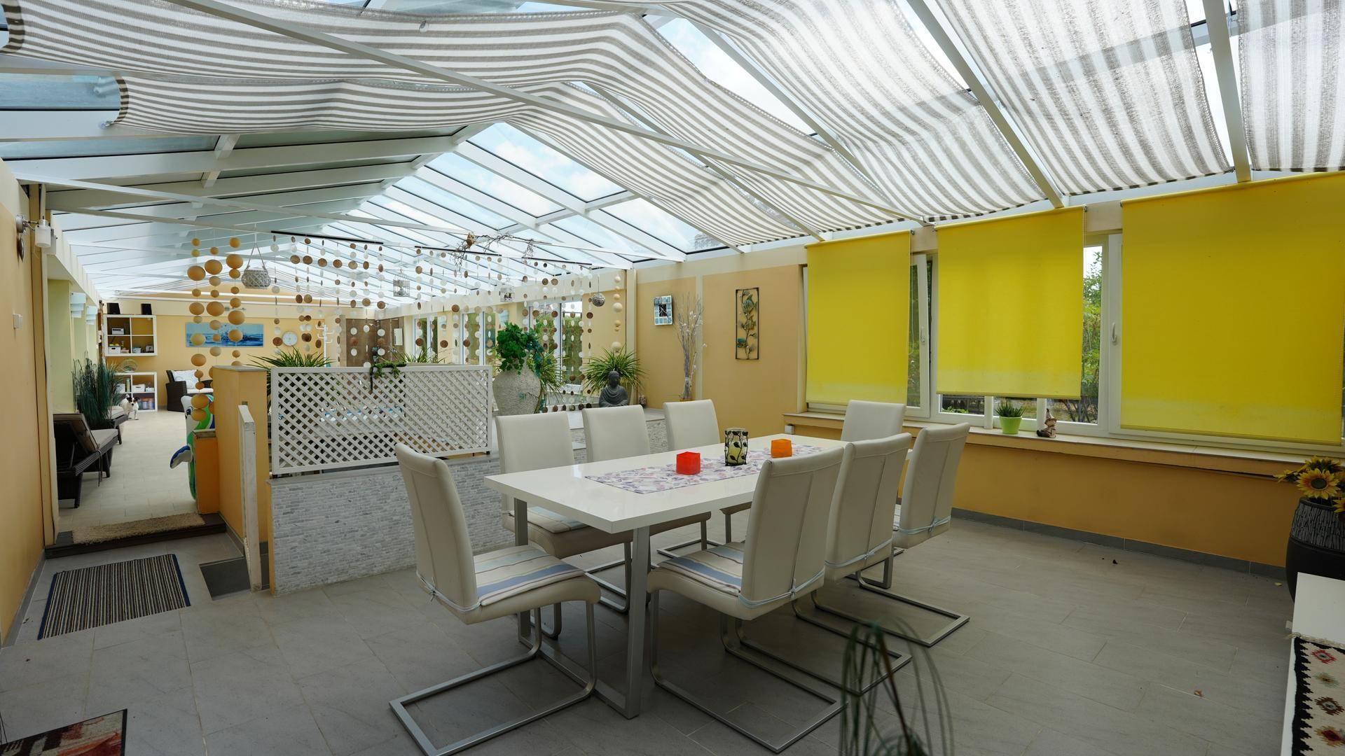 Heller Essbereich im Wintergarten mit großem Tisch, modernen Stühlen und gestreiften Sonnenschutzrollos.