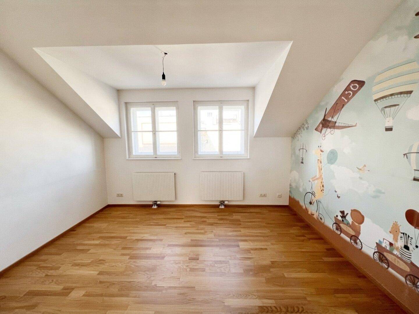 Geräumiges Kinderzimmer mit Holzboden, zwei Fenstern und einer charmanten Wanddekoration.