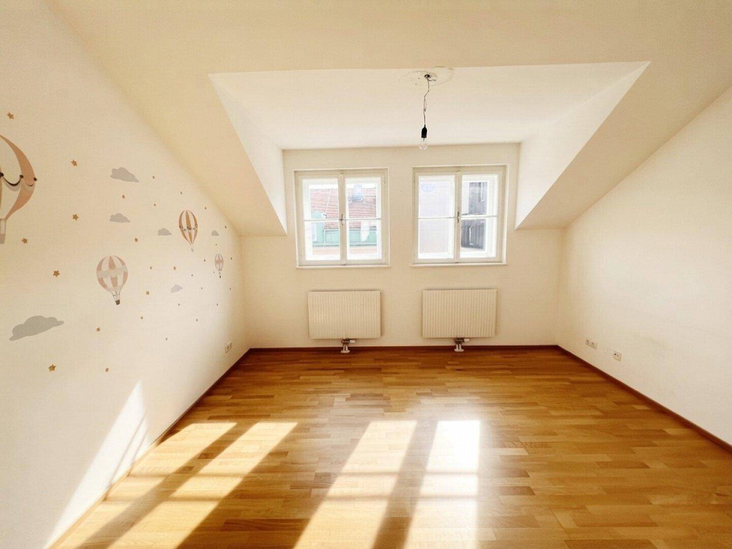Freundliches Zimmer mit Holzboden und zwei Fenstern, das viel Tageslicht hereinlässt.