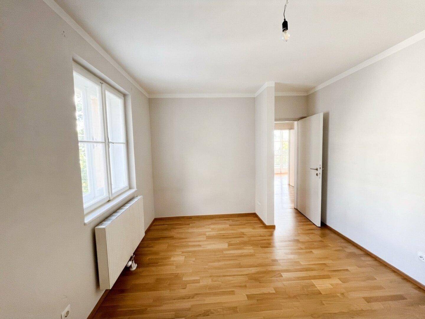 Leeres Schlafzimmer mit Parkettboden, weißen Wänden und großem Fenster.