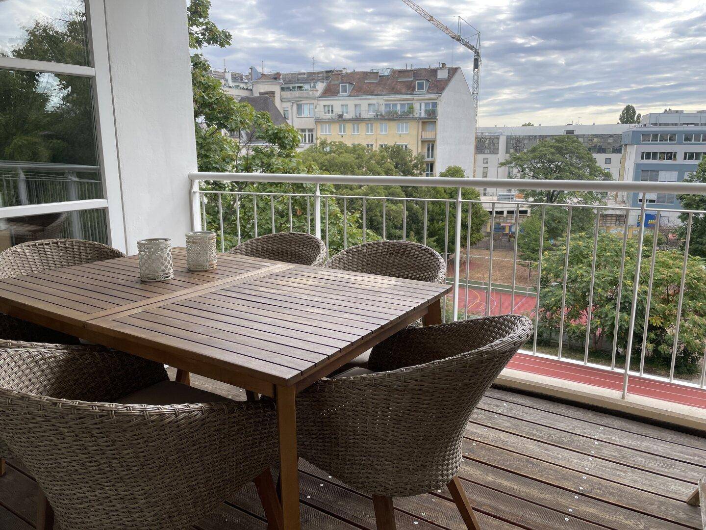 Möblierter Balkon mit Holztisch und Stühlen, Blick auf Sportplatz und Stadt.