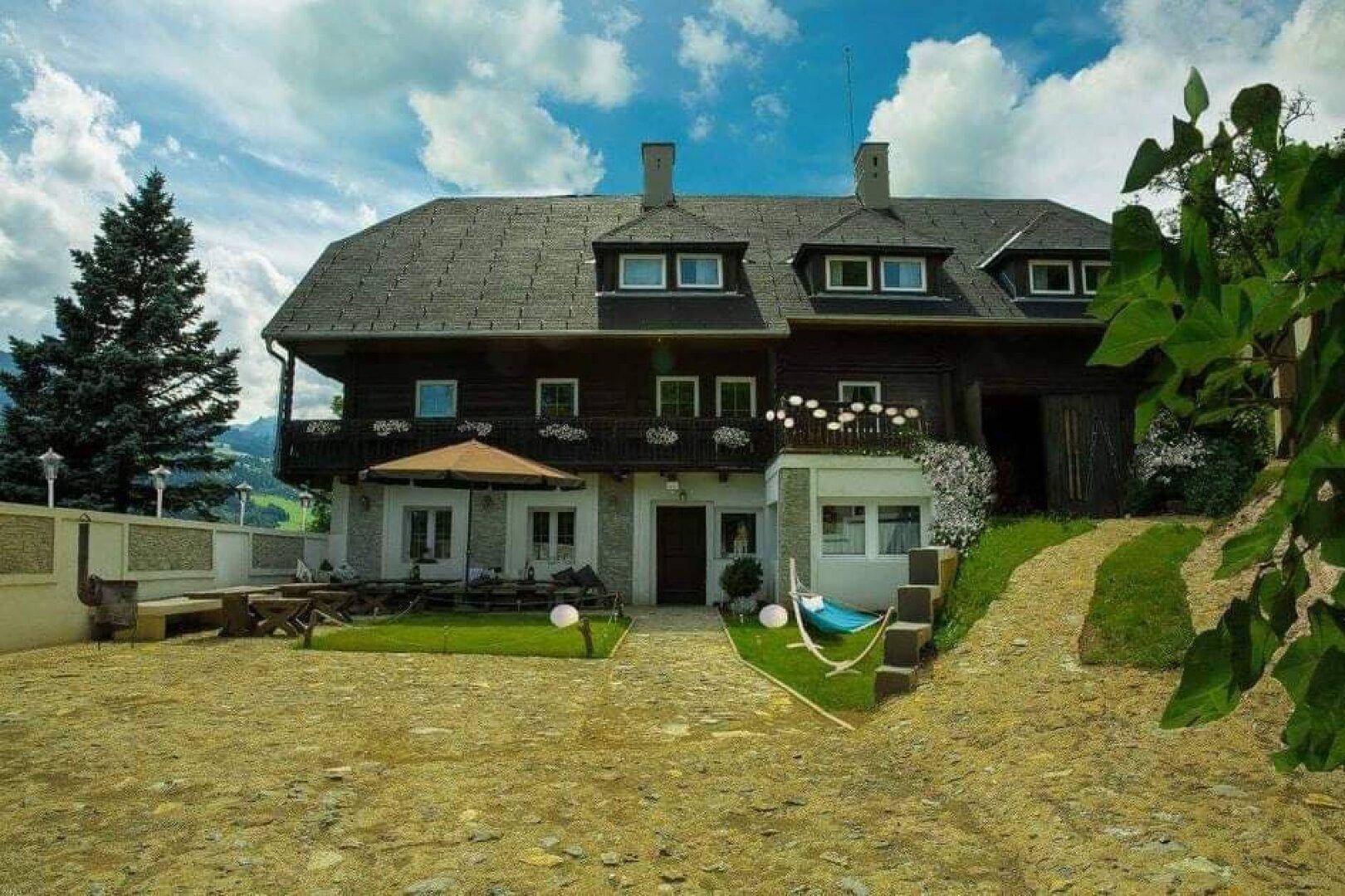 Renoviertes Gebäude mit gemütlicher Terrasse, Sonnenschirm und Hängematte vor einer grünen Berglandschaft.