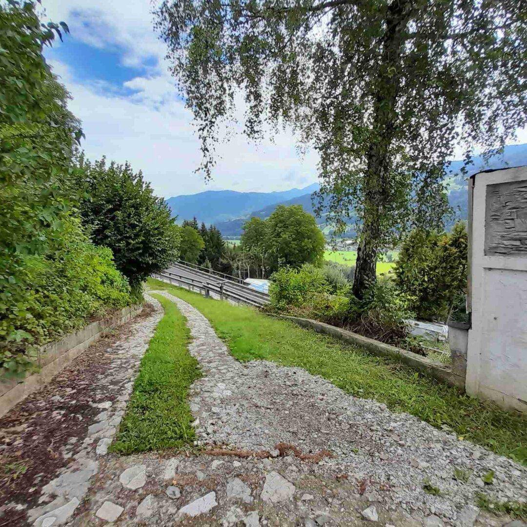 Steiniger Weg, der zu einem Gebäude führt, umgeben von Bäumen und einer weiten grünen Landschaft.