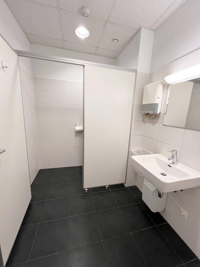 Saubere und funktionale Toilette mit weißen Fliesen, dunklem Boden und einem Waschbecken, ausgestattet mit Spender.