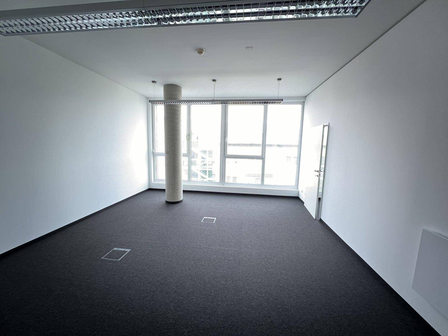 Modernes Büro mit einer Betonsäule, großen Fenstern und Teppichboden, ideal für eine flexible Büronutzung.