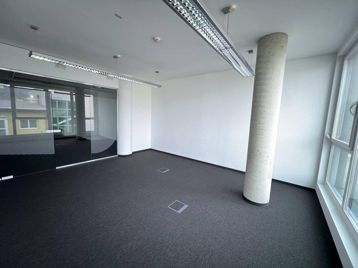 Heller Büroraum mit Teppichboden, einer Betonsäule und einer modernen Glaswand, die einen separaten Bereich abtrennt.