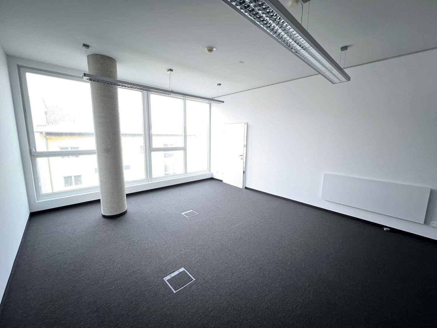 Geräumiges Büro mit Teppichboden, einer Betonsäule und großen Fenstern, die viel Licht in den Raum lassen.