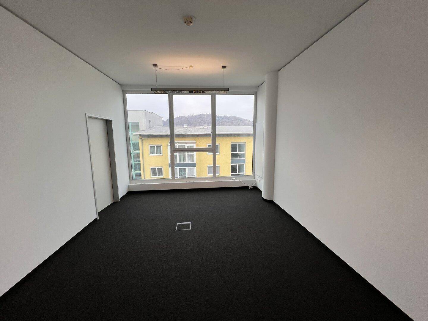Kompaktes Büro mit Teppichboden und einem großen Fenster, das einen Blick auf die gegenüberliegenden Gebäude bietet.
