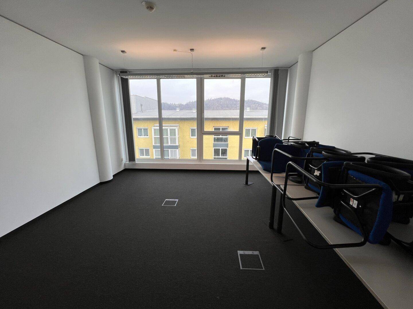 Büroraum mit Teppichboden und großen Fenstern, die einen Blick auf die umliegende Bebauung ermöglichen, teilweise möbliert.