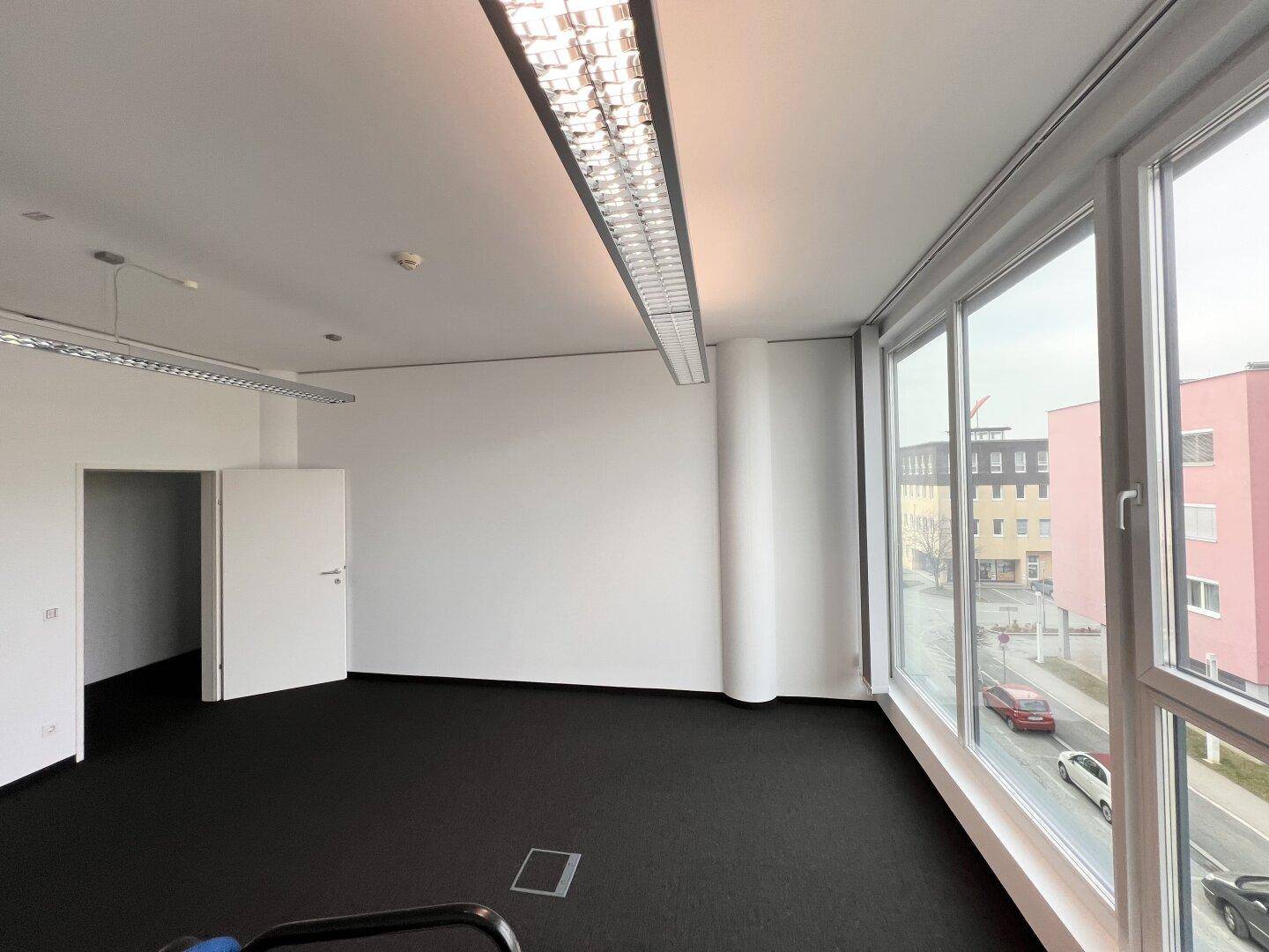 Helles Eckbüro mit Teppichboden und durchgehenden Fenstern, die einen weiten Blick auf die Stadt bieten.