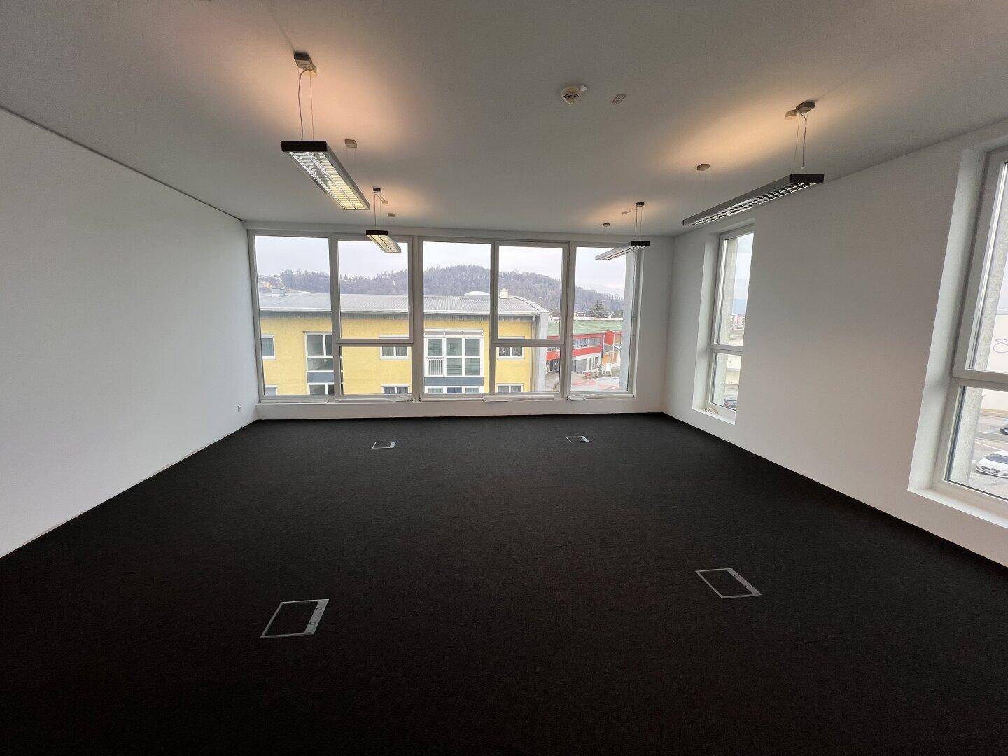 Helles und modernes Büro mit Teppichboden und großen Fenstern, die viel Tageslicht hereinlassen.