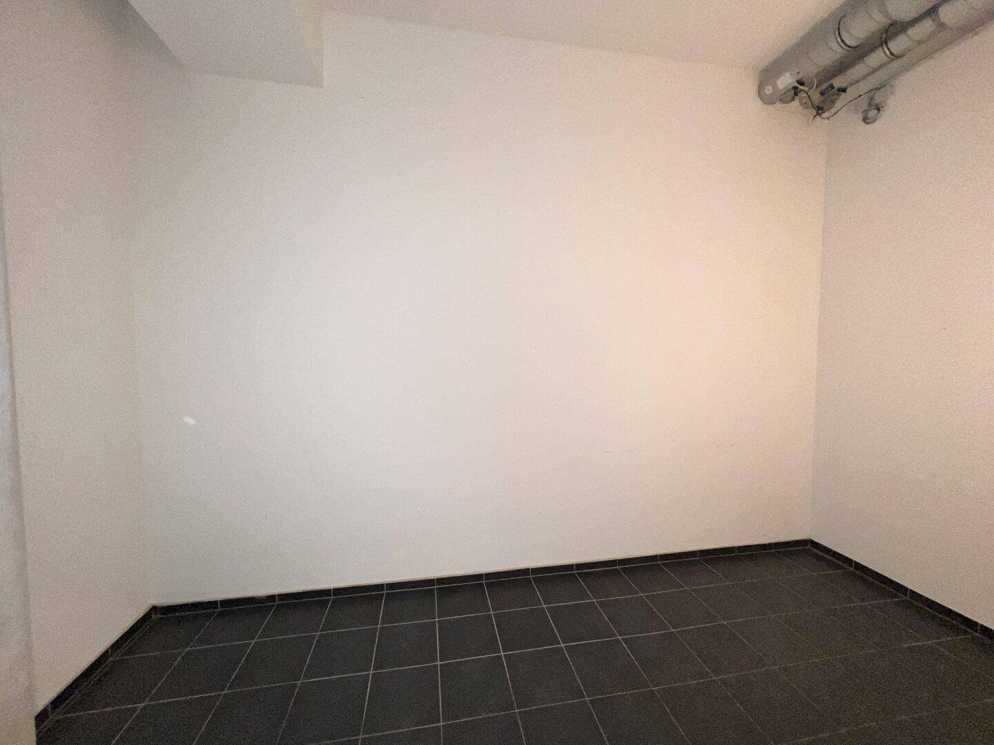 Kleiner, leerer Raum mit dunklem Fliesenboden und sichtbaren Lüftungsrohren an der Decke, ideal als Lager.