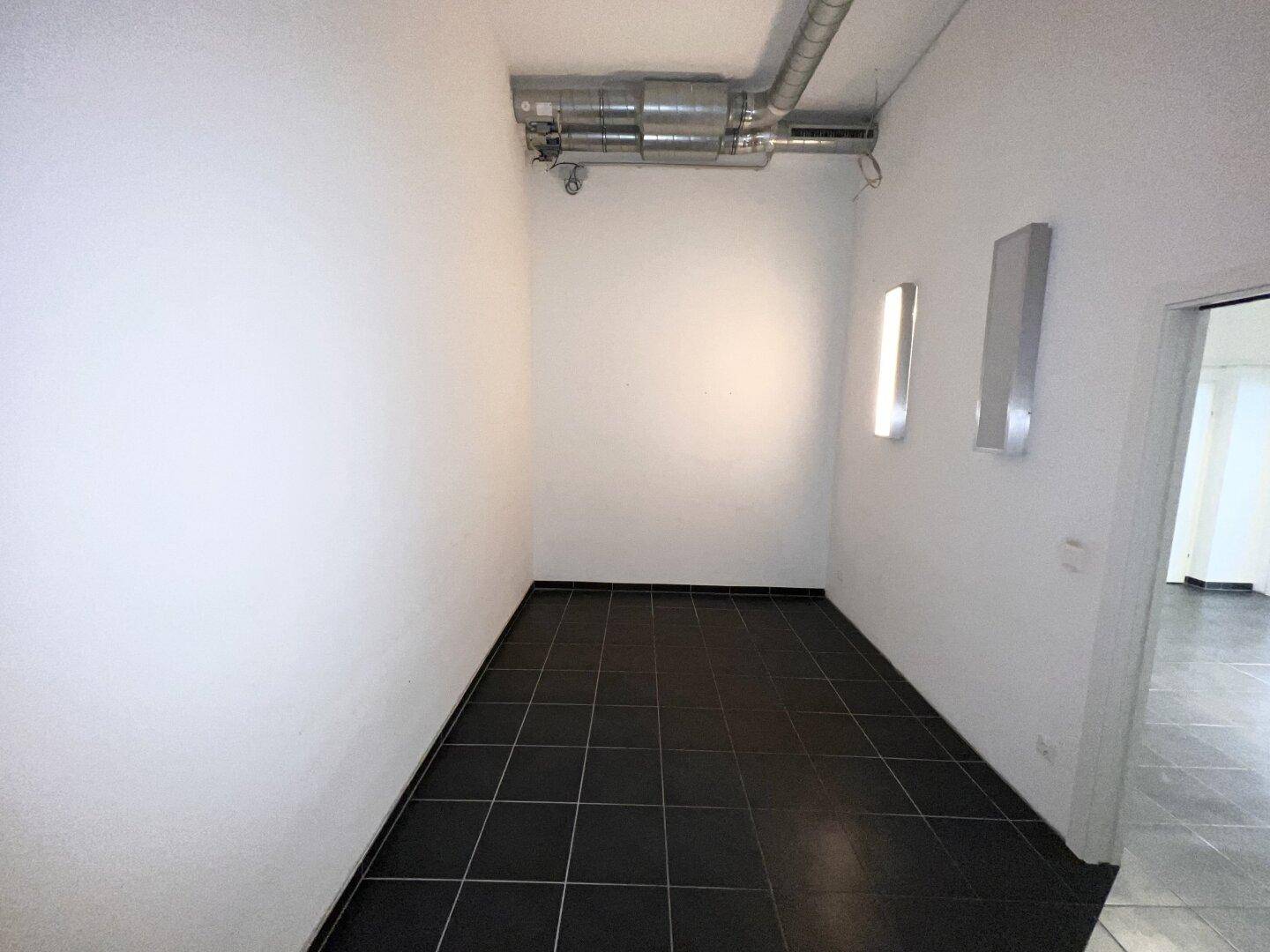 Leerer Raum mit dunklem Fliesenboden, weißen Wänden und Deckenbeleuchtung, geeignet für verschiedene Zwecke.