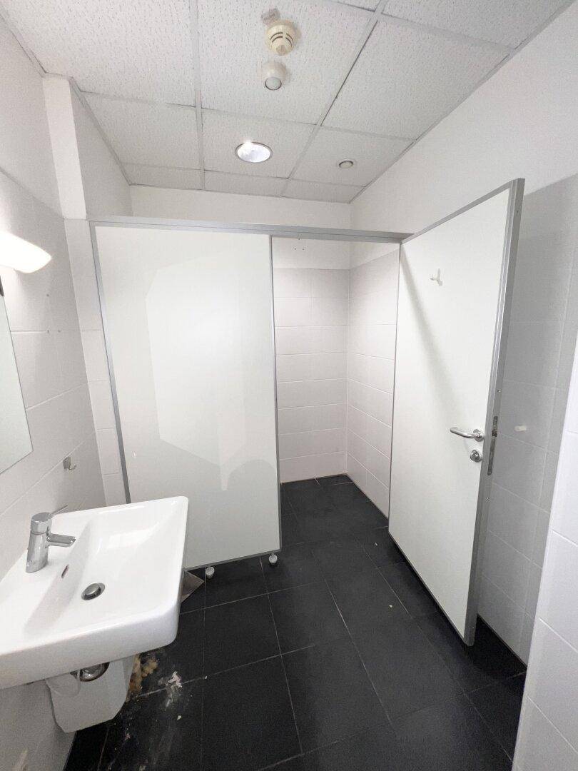 Moderner Toilettenbereich mit Waschbecken, Spiegel und weißen Trennwänden in einem Gewerbeobjekt.