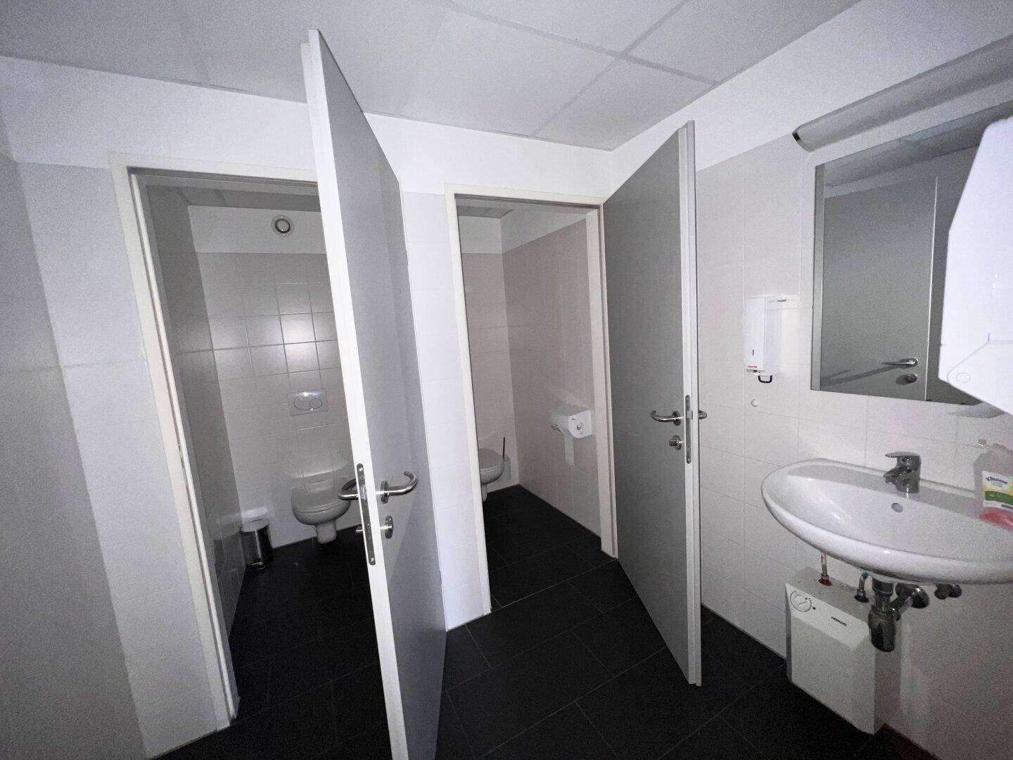 Waschraum mit mehreren Toilettenkabinen, Waschbecken und Spiegel.