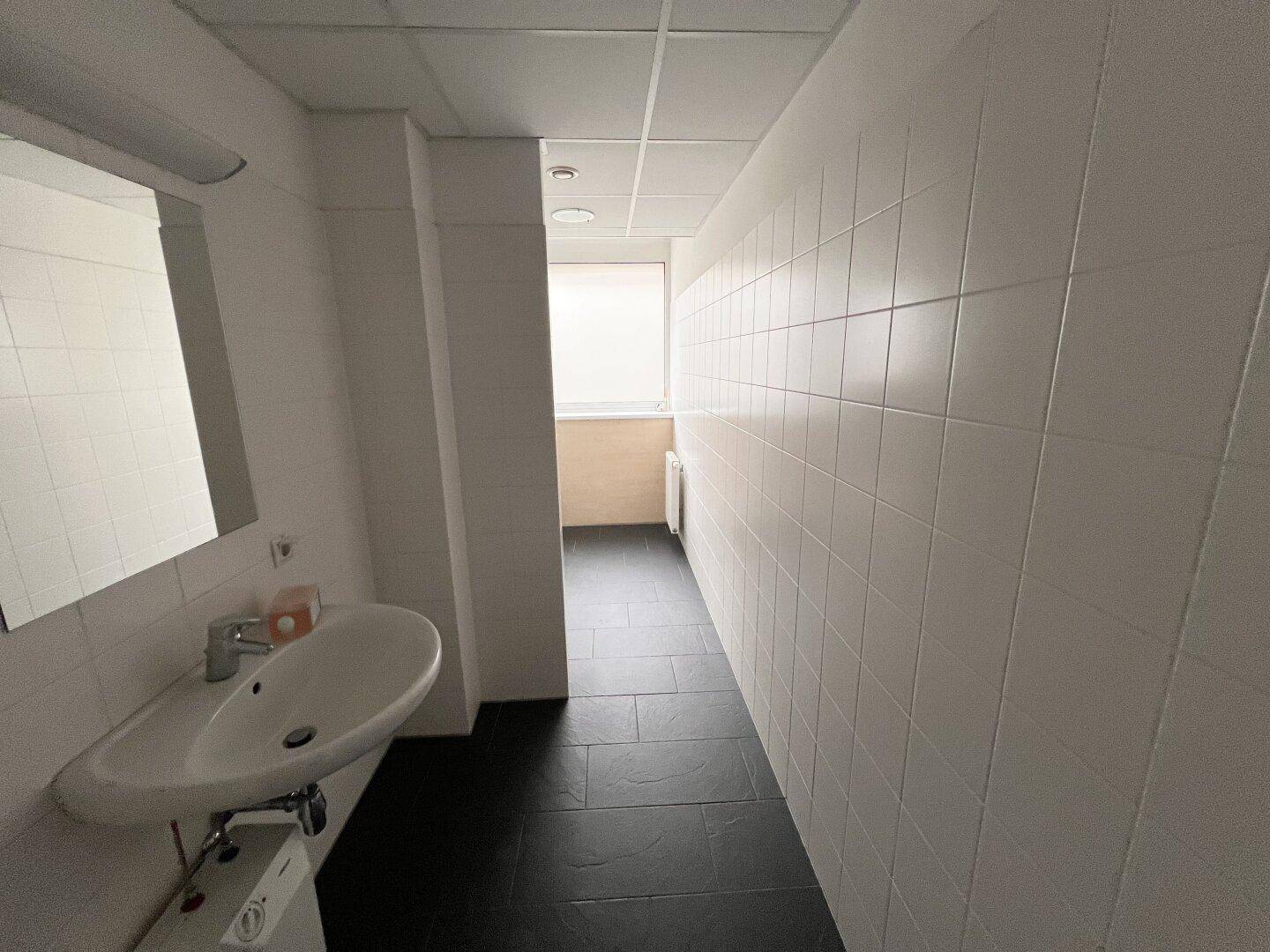 Langer Waschraum mit weißen Fliesenwänden, dunklem Boden und einem Waschbecken.