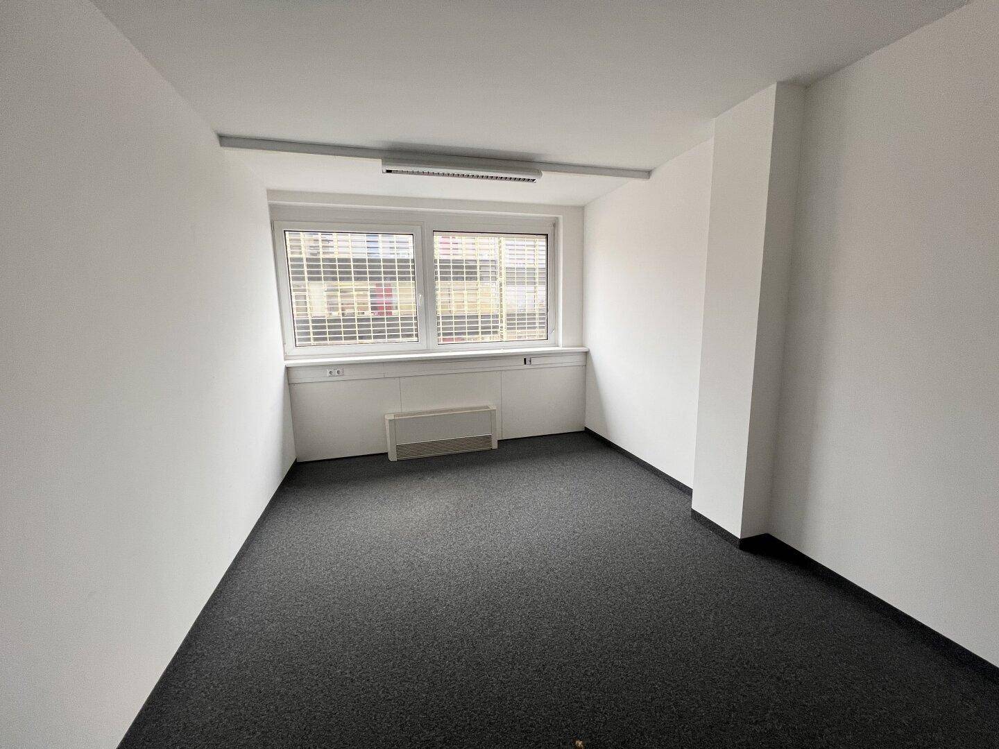 Kleineres Büro mit Teppichboden, einem Fenster mit Jalousien und weiß gestrichenen Wänden.