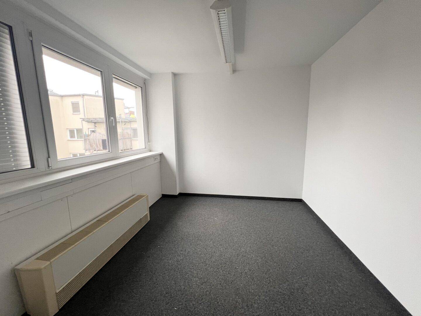 Leeres Büro mit Teppichboden, einem Fenster mit Jalousien und einem Heizkörper unter dem Fenster.