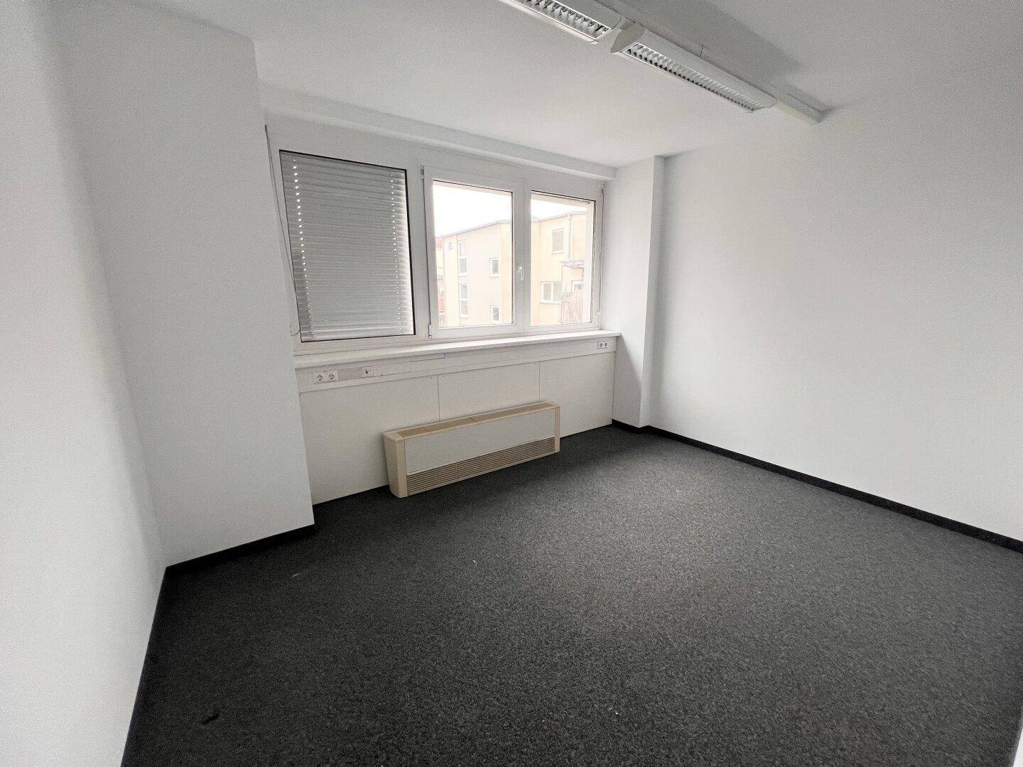 Helles Büro mit Teppichboden, breiter Fensterfront mit Jalousien und weißen Wänden.